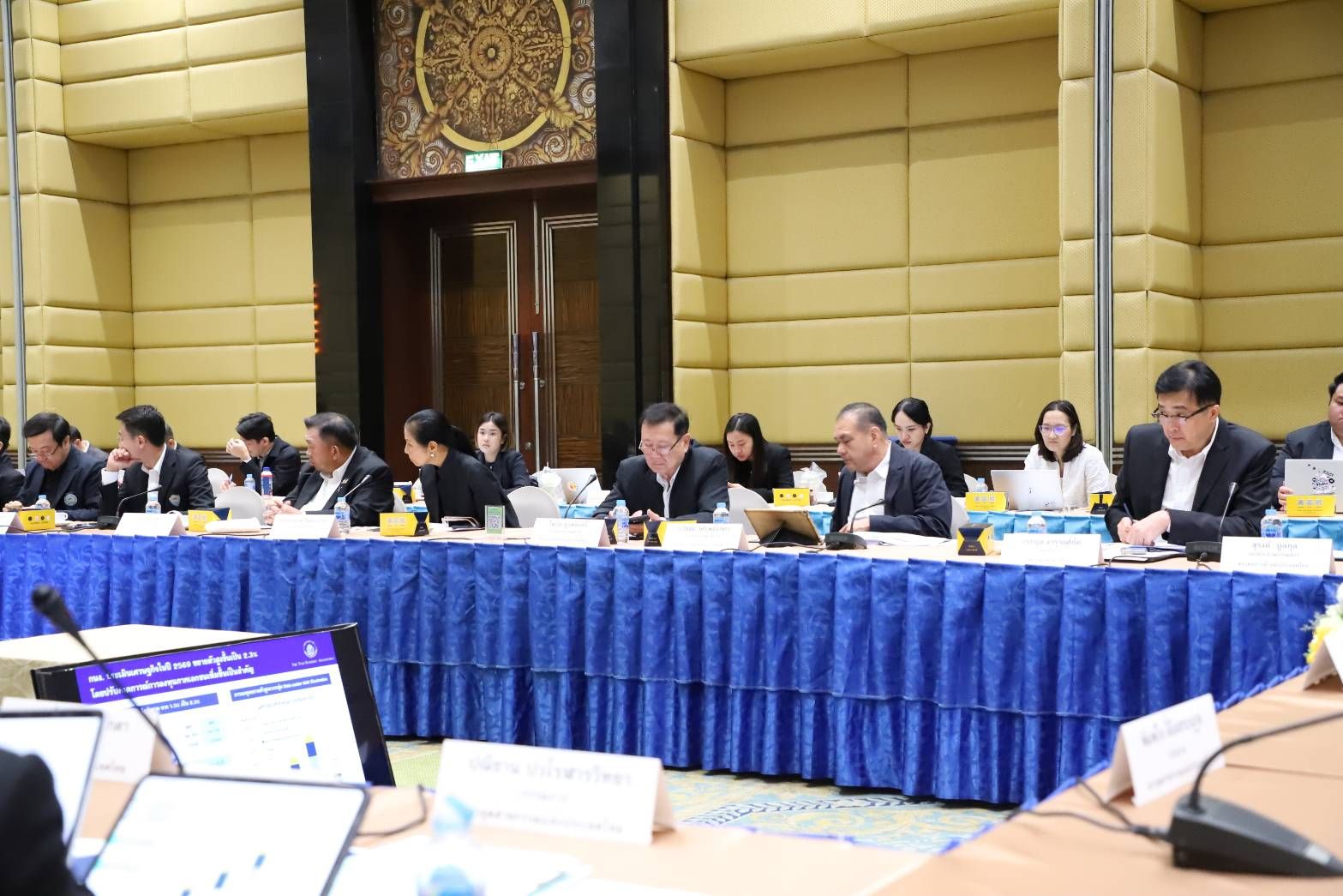 Professionals attend a formal meeting, seated at a long table with a blue tablecloth.
