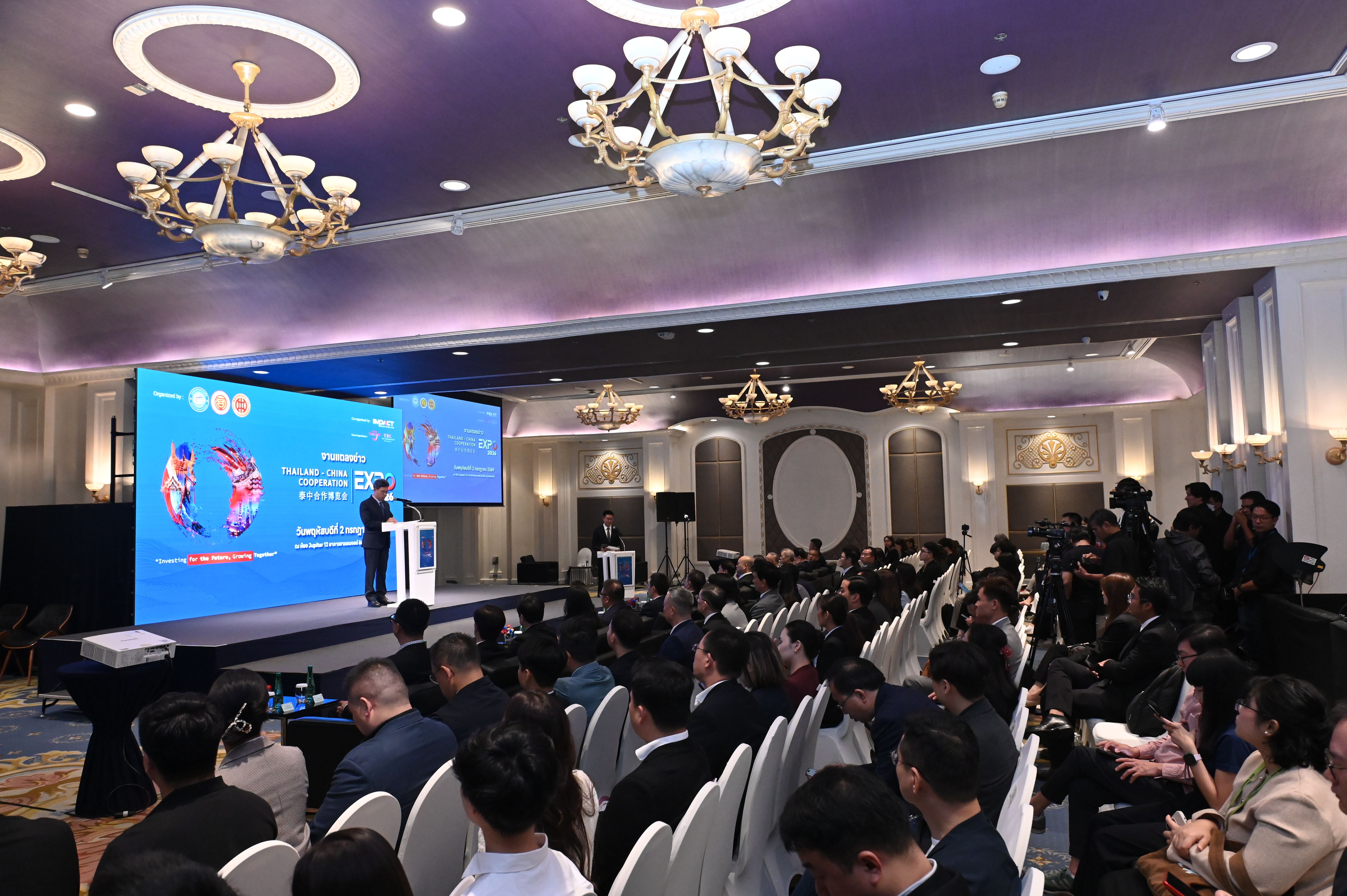 A speaker presents to a large audience in a decorated ballroom with a bright screen.