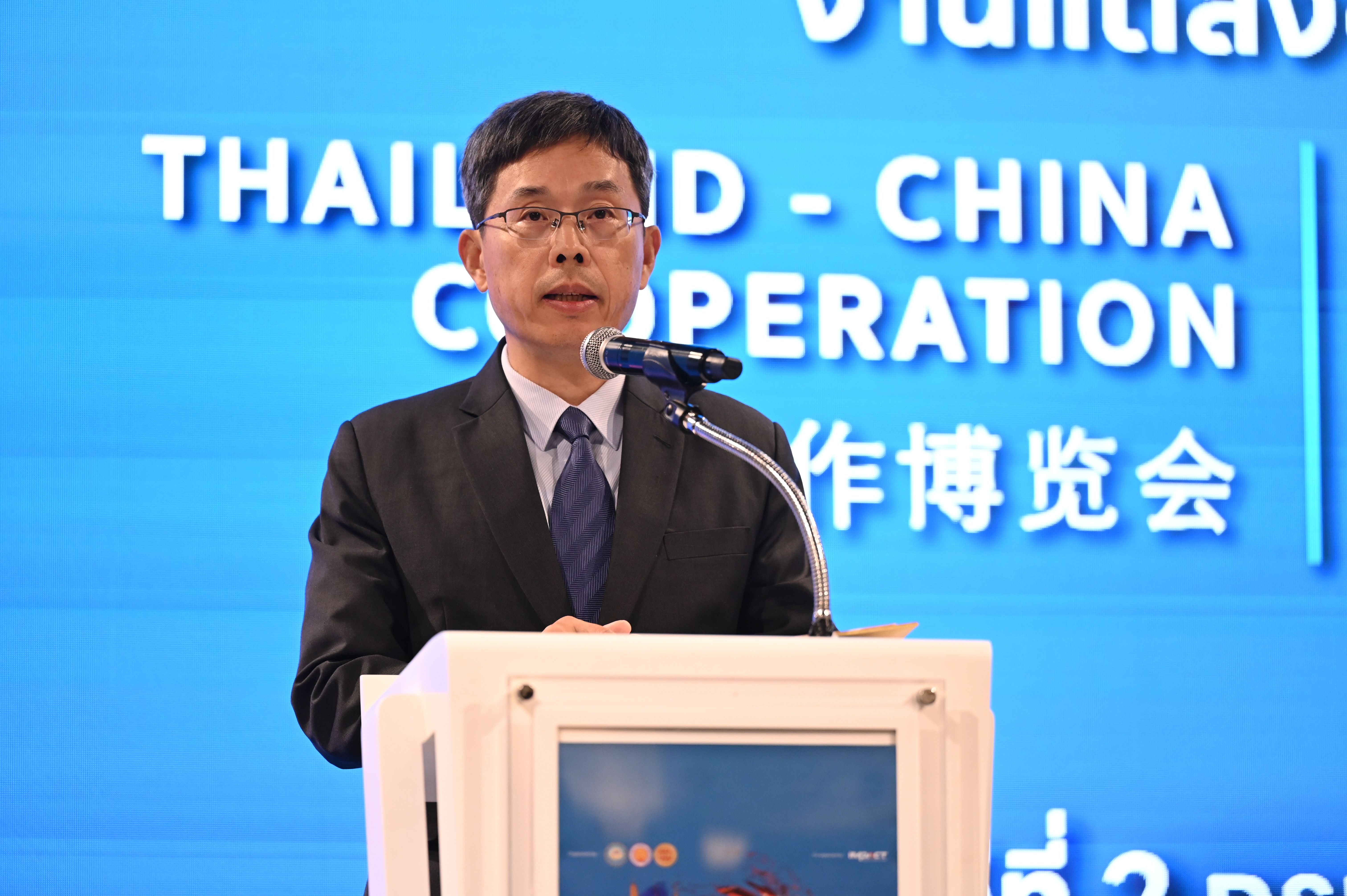 Asian man in glasses and suit speaks at podium during Thailand-China cooperation event.