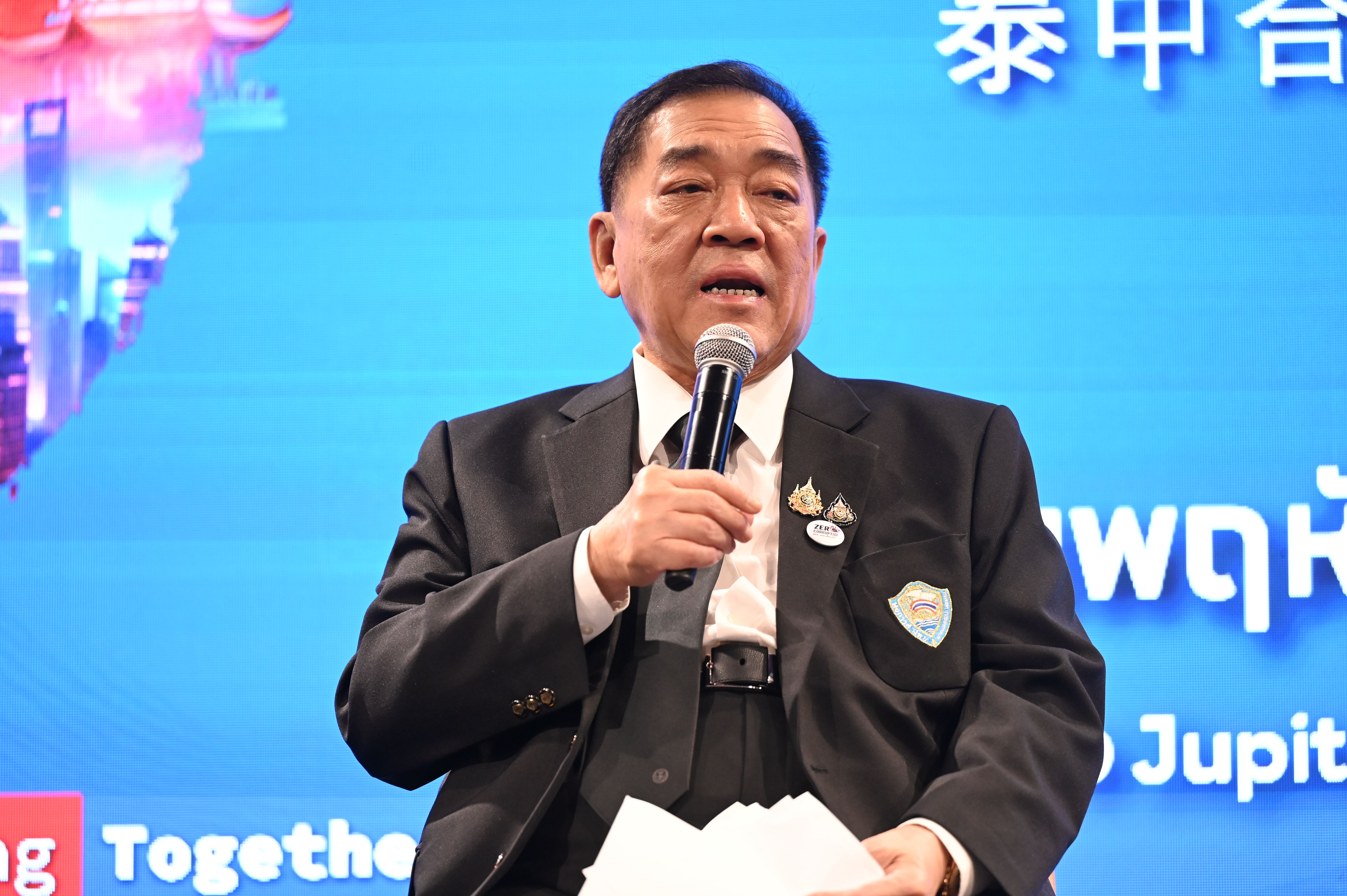 Older Asian man in a dark suit speaking into microphone, holding papers, blue backdrop.