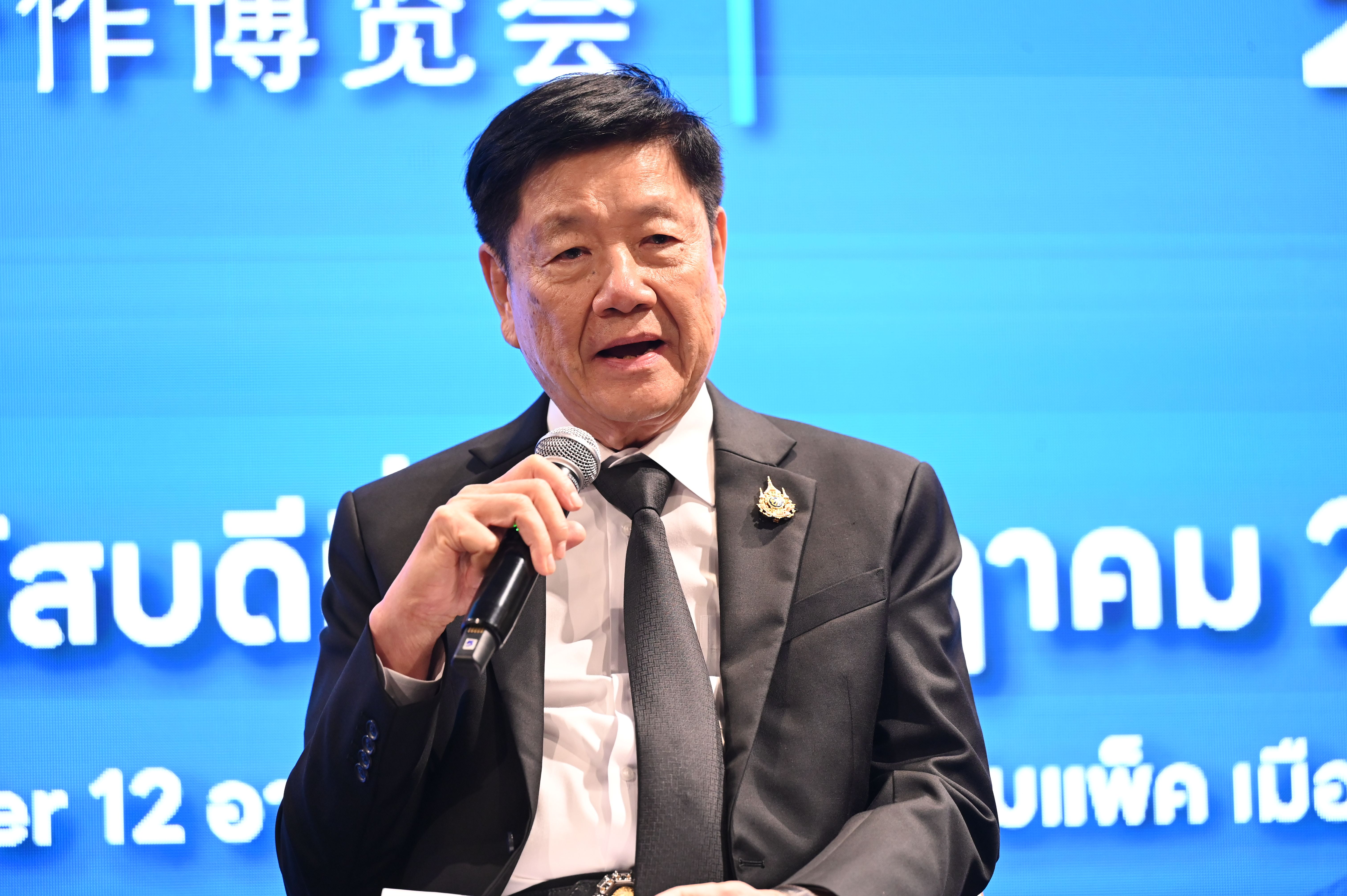 A man in a suit speaks into a microphone against a blue background with text.