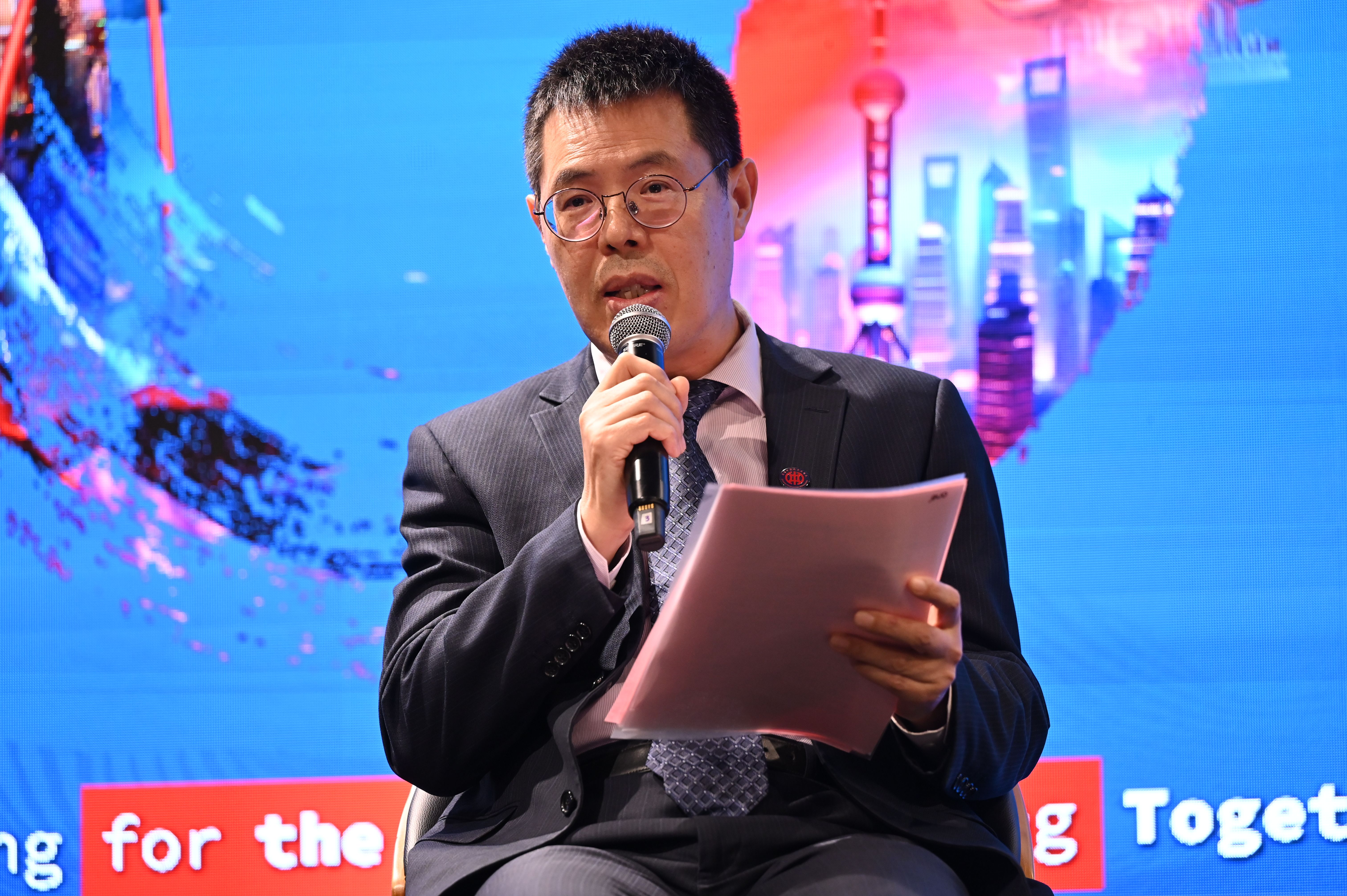 A man in a dark suit speaks into a microphone while holding documents at an event.
