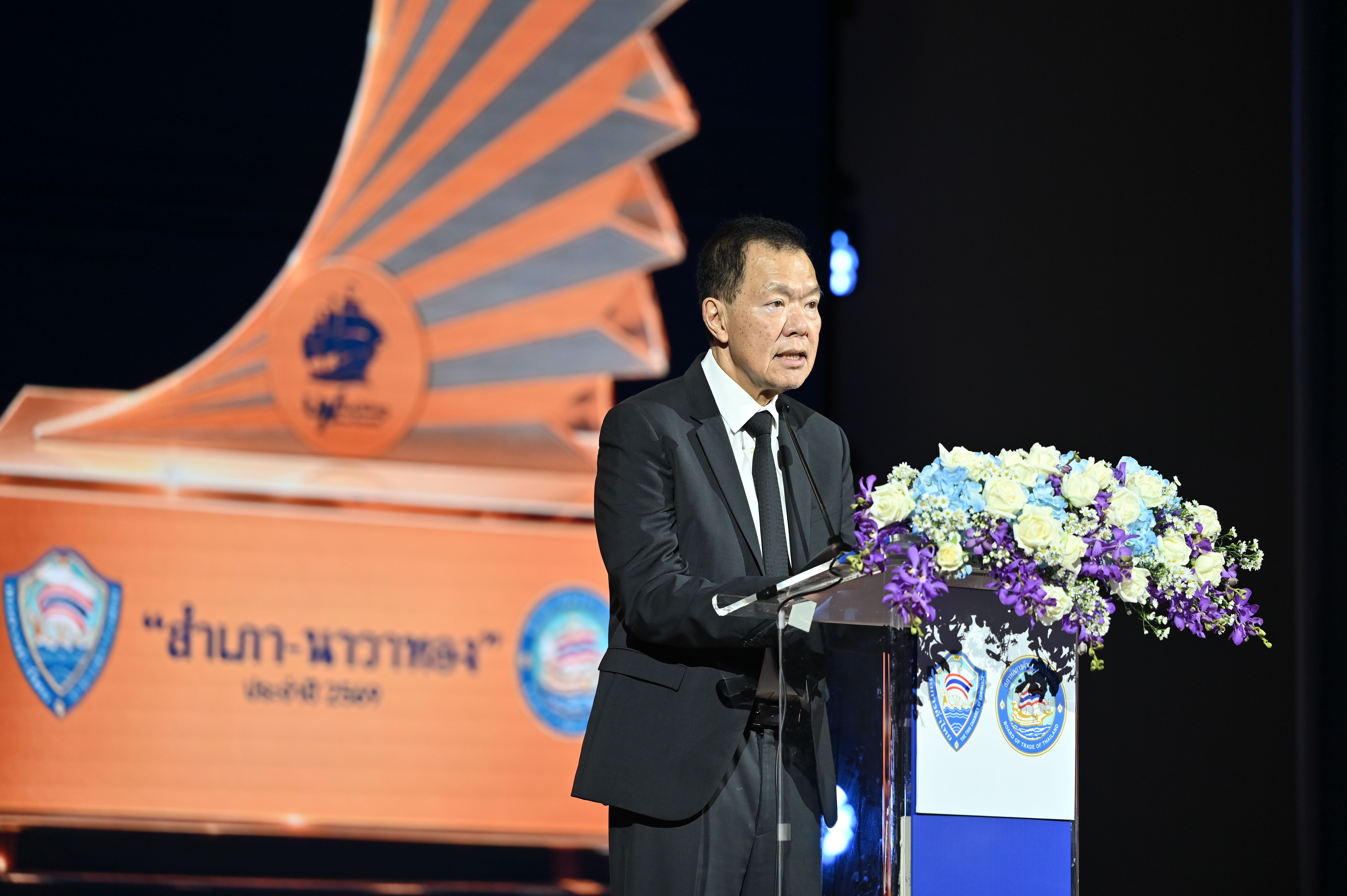 Man in a dark suit speaking at a podium with flowers and an elaborate golden backdrop.
