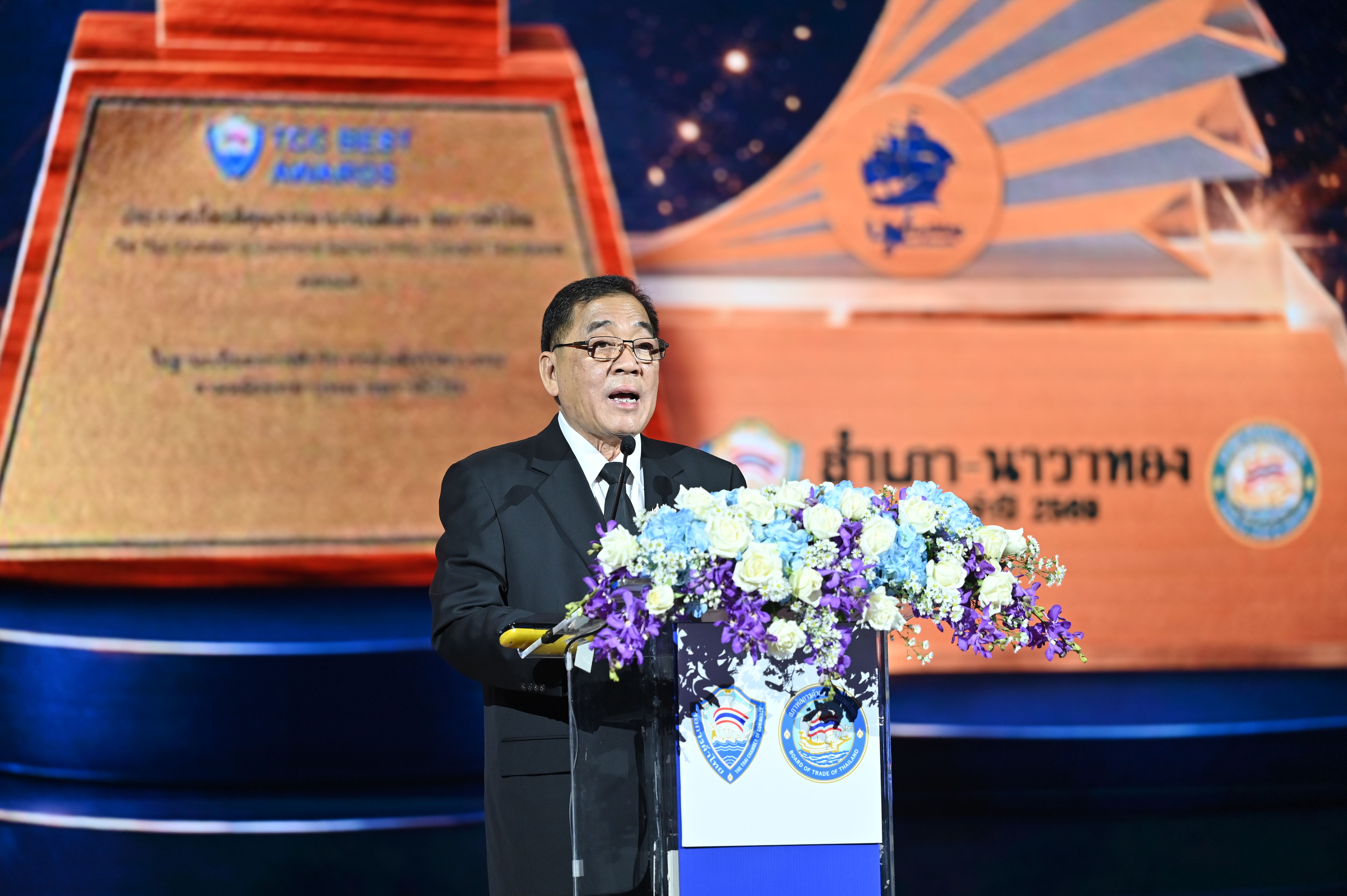 Man in suit speaks at podium with flowers, an award plaque visible in background.