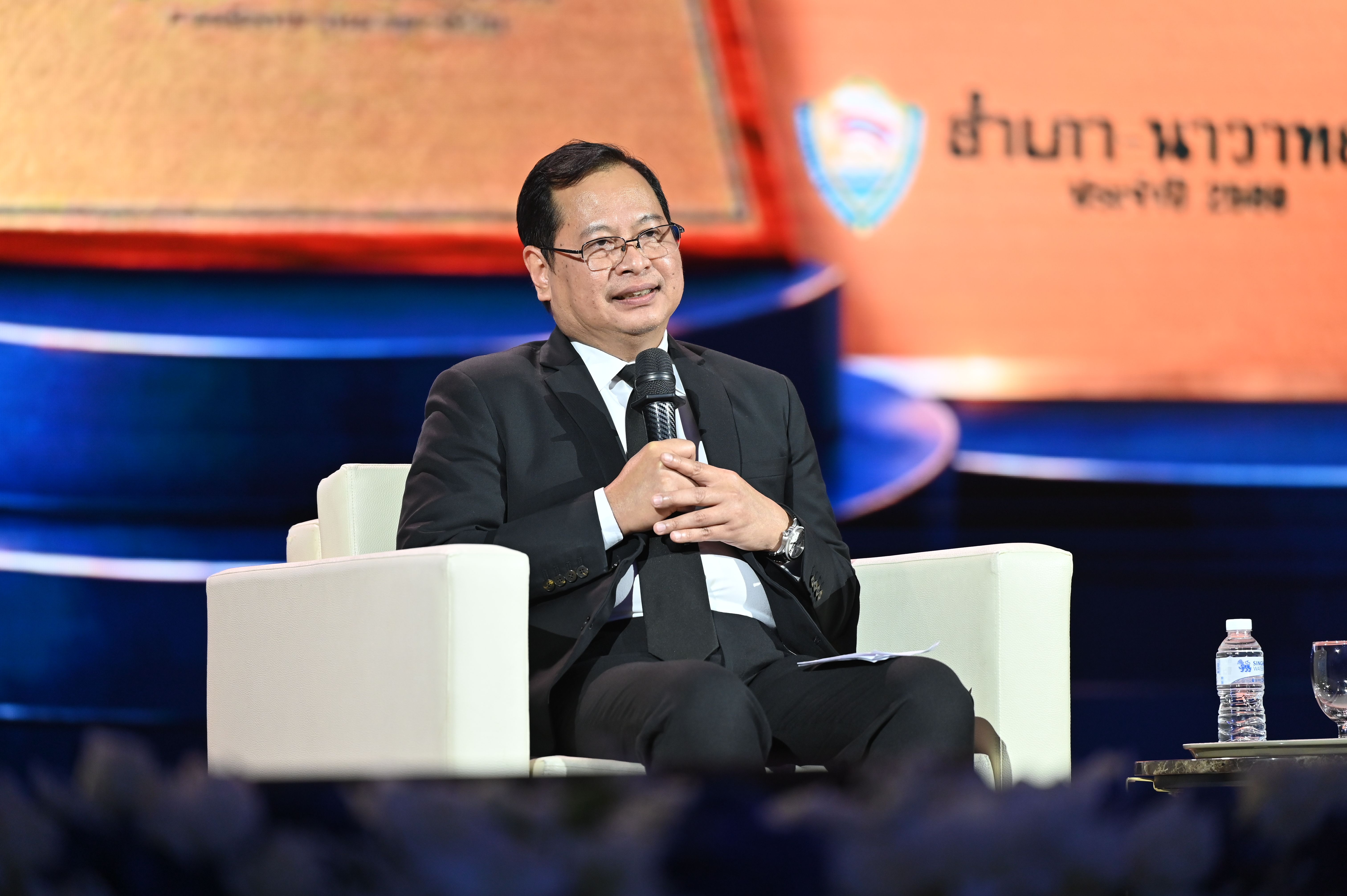 Man in suit and glasses speaking into a microphone on a stage.