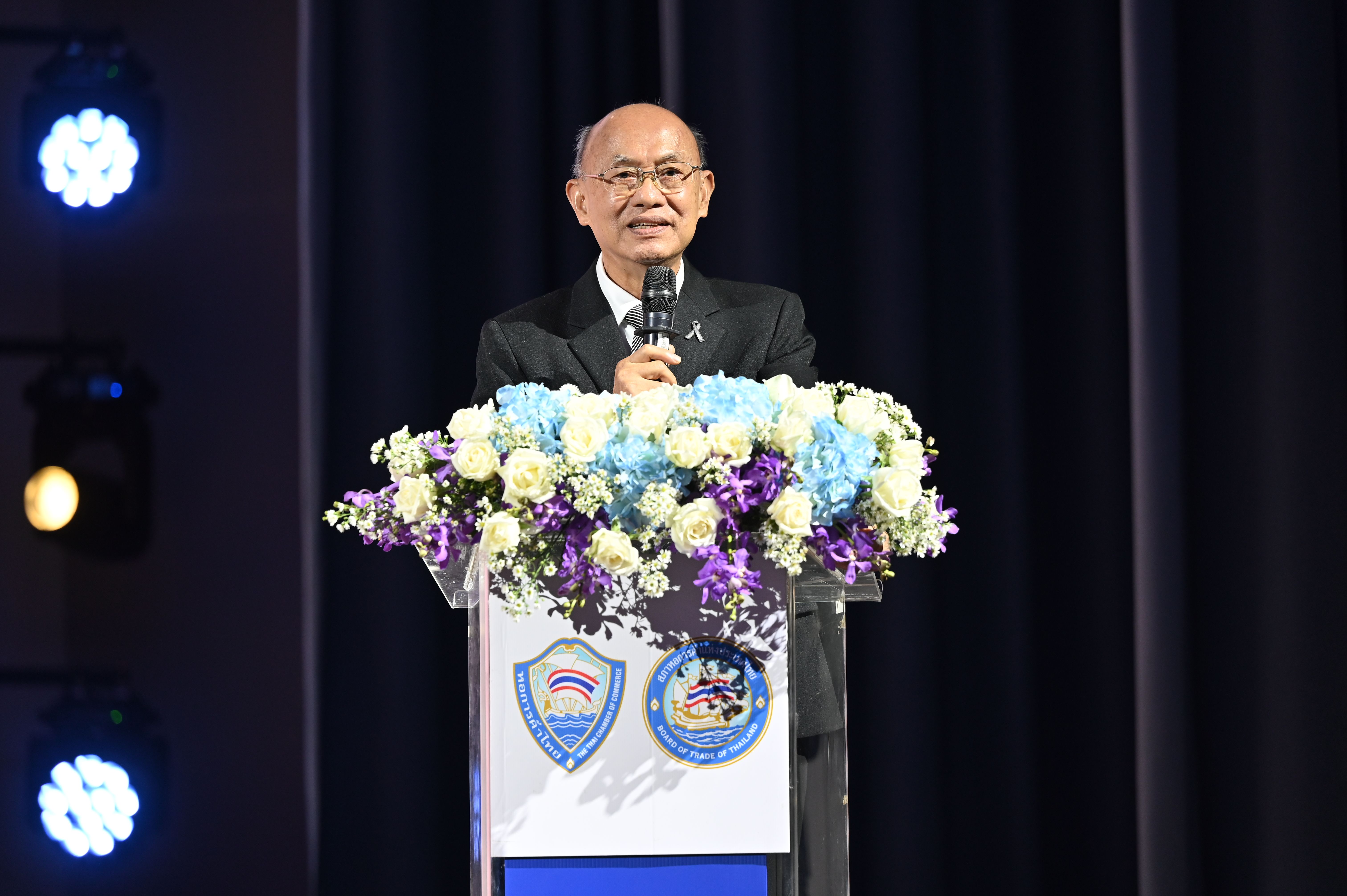 A man in a dark suit speaks at a podium adorned with flowers, holding a microphone.