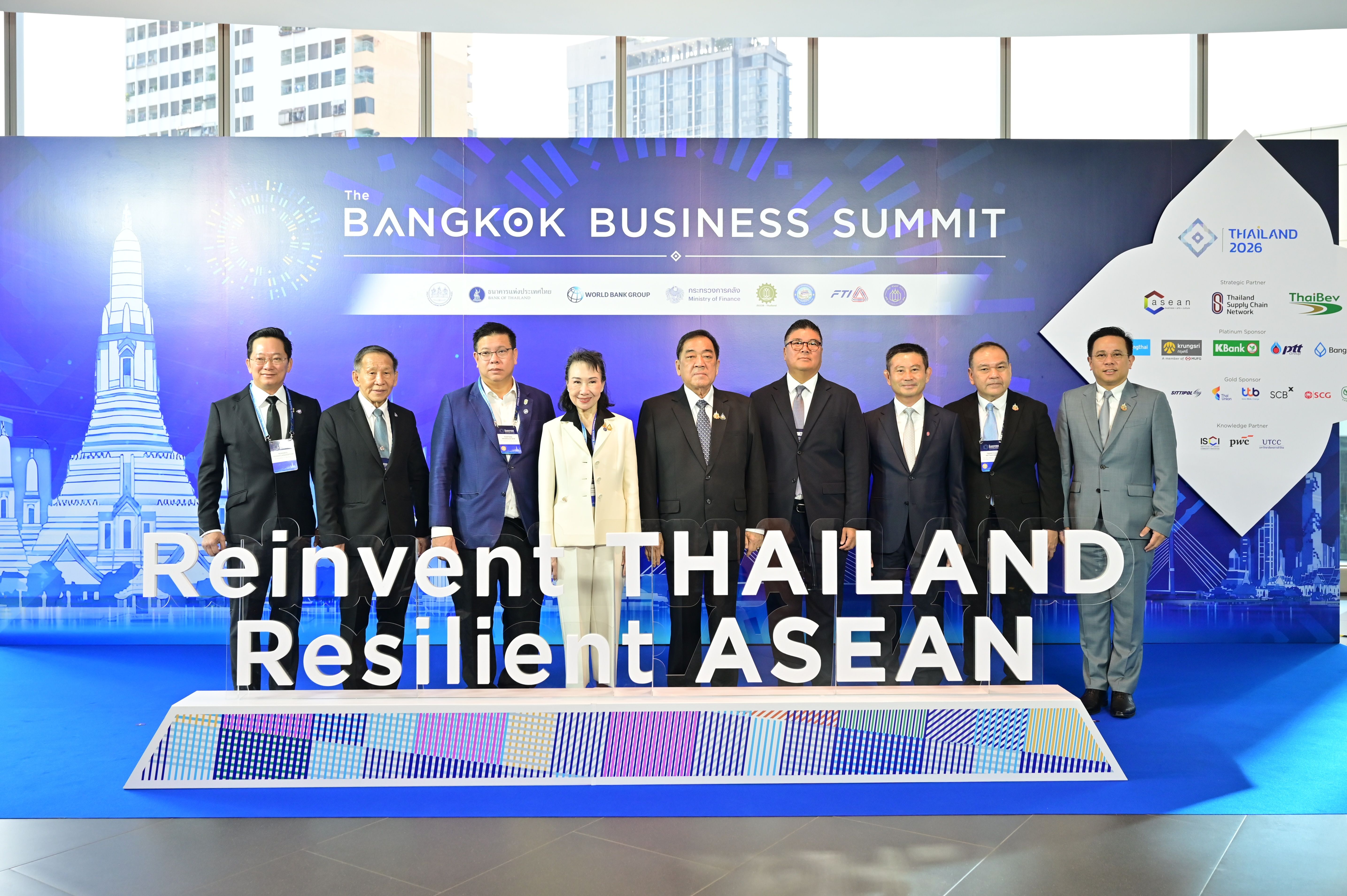 Nine professionals pose at the Bangkok Business Summit with the "Reinvent THAILAND Resilient ASEAN" banner.