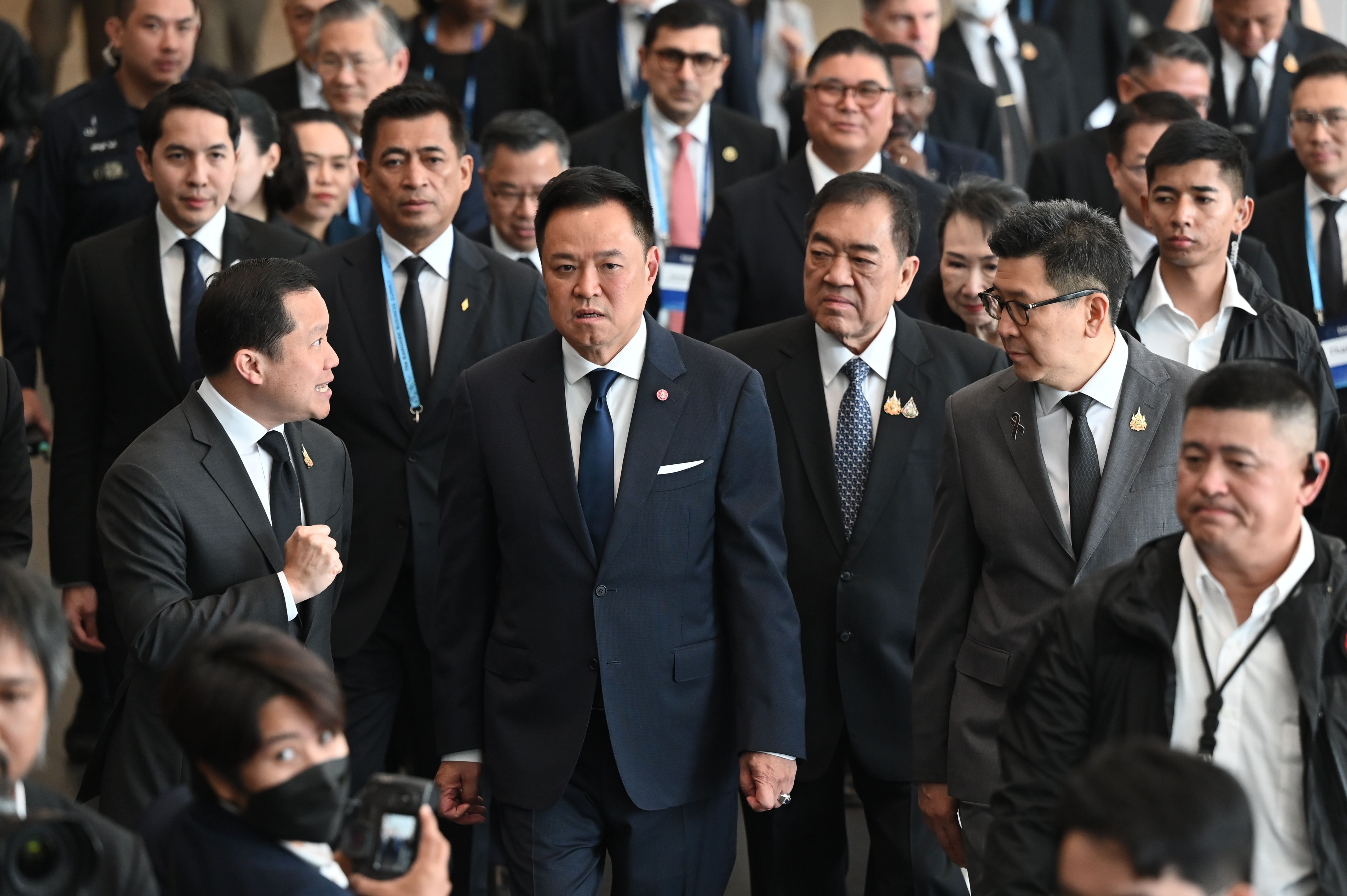 A man in a dark suit walks forward, surrounded by other men in business attire at an event.
