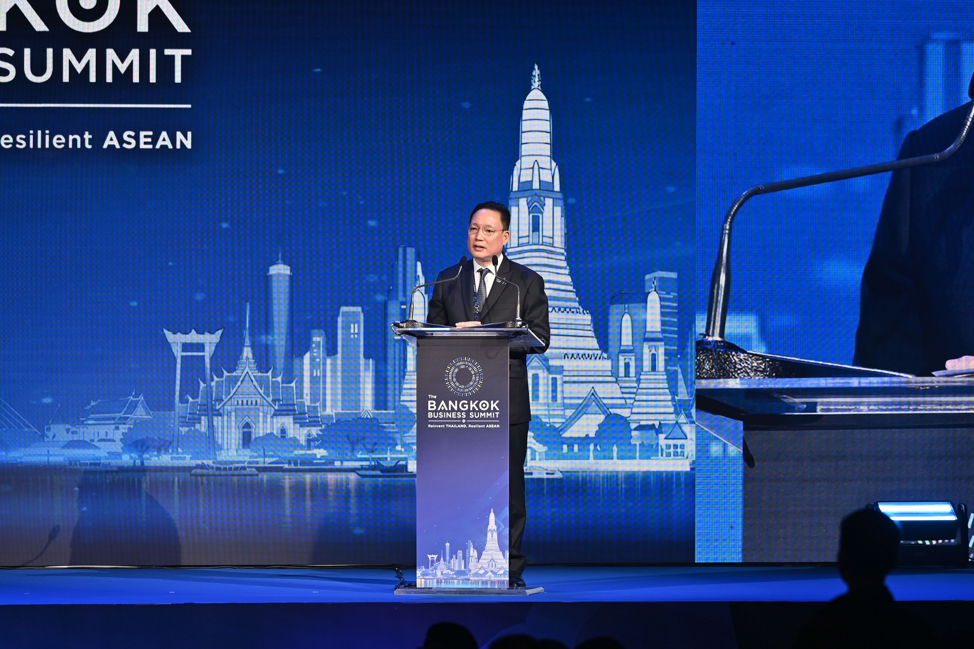 Man speaks at podium before large screen displaying Bangkok skyline at an international summit.