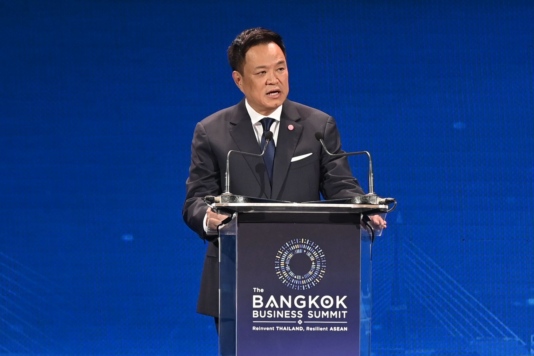Man in dark suit speaks at podium with 'The Bangkok Business Summit' logo.
