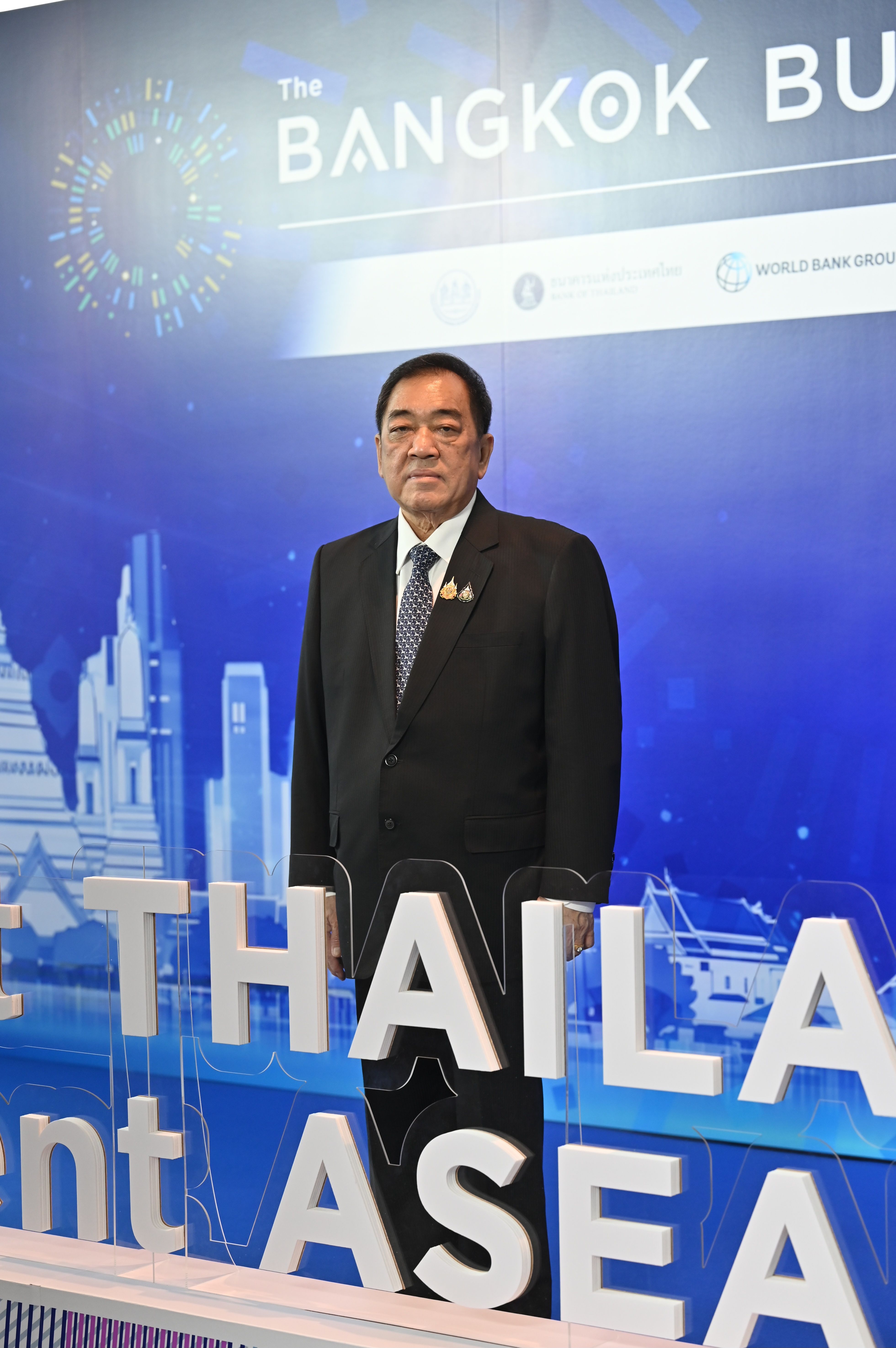 A man in a dark suit and tie stands before a blue banner with 'Thailand ASEAN'.