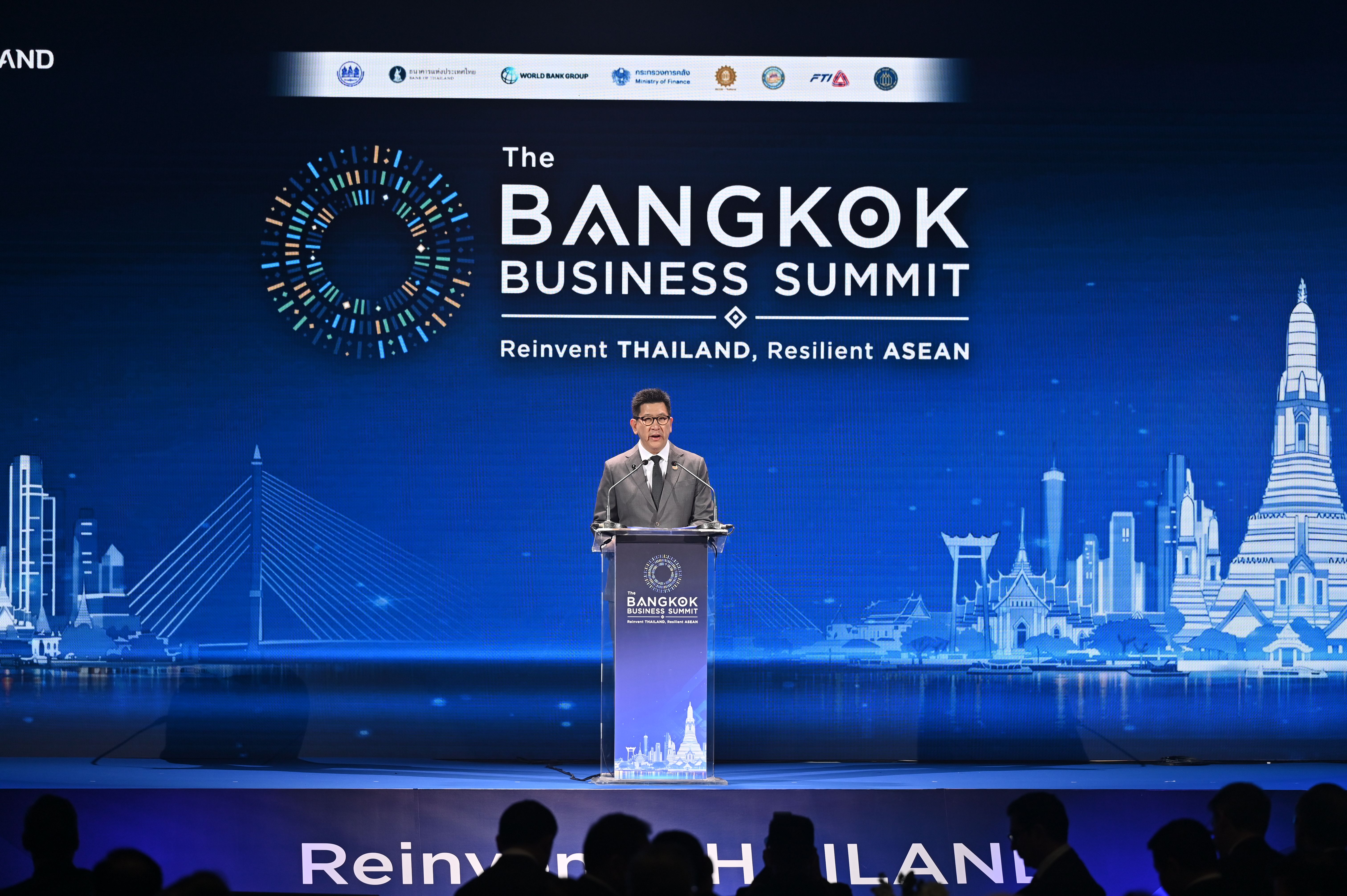 Man speaking at podium during The Bangkok Business Summit with city skyline backdrop.
