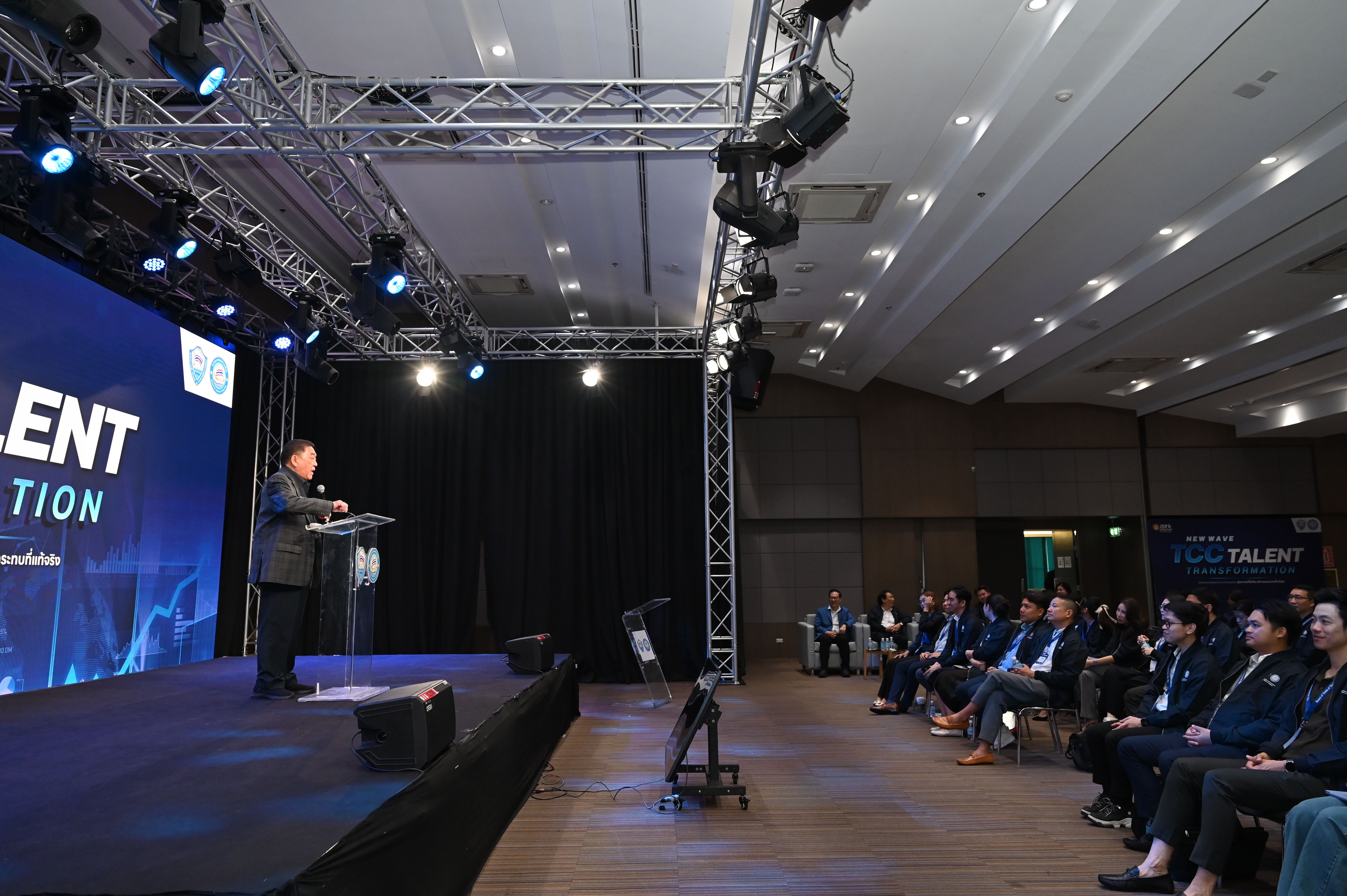 A man speaks on a stage with a large screen displaying 'TALENT' to an audience.