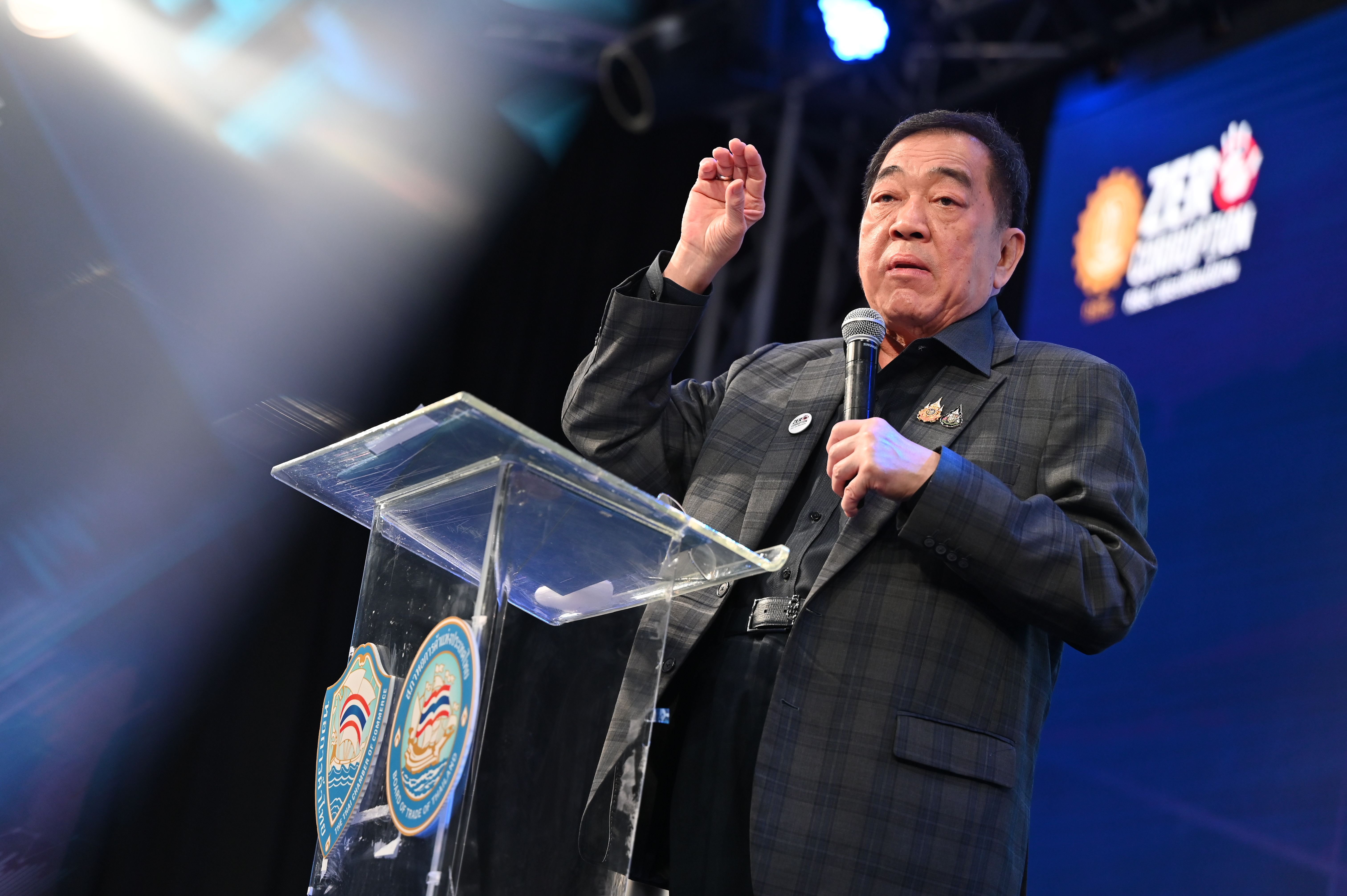 Asian man in plaid suit speaks animatedly at a clear podium, hand raised.