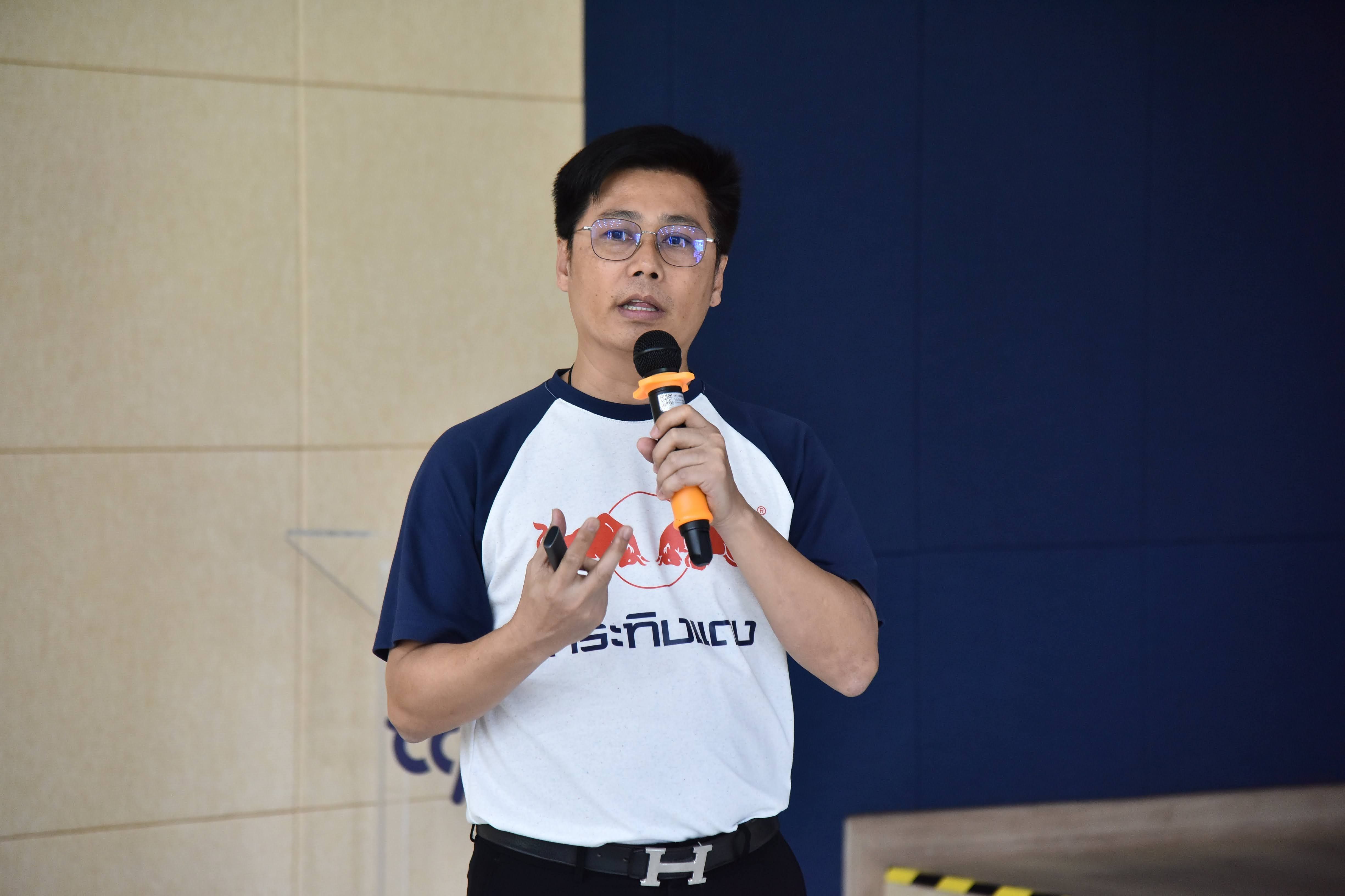 A man in glasses speaks into a microphone, gesturing with his hands during a presentation.