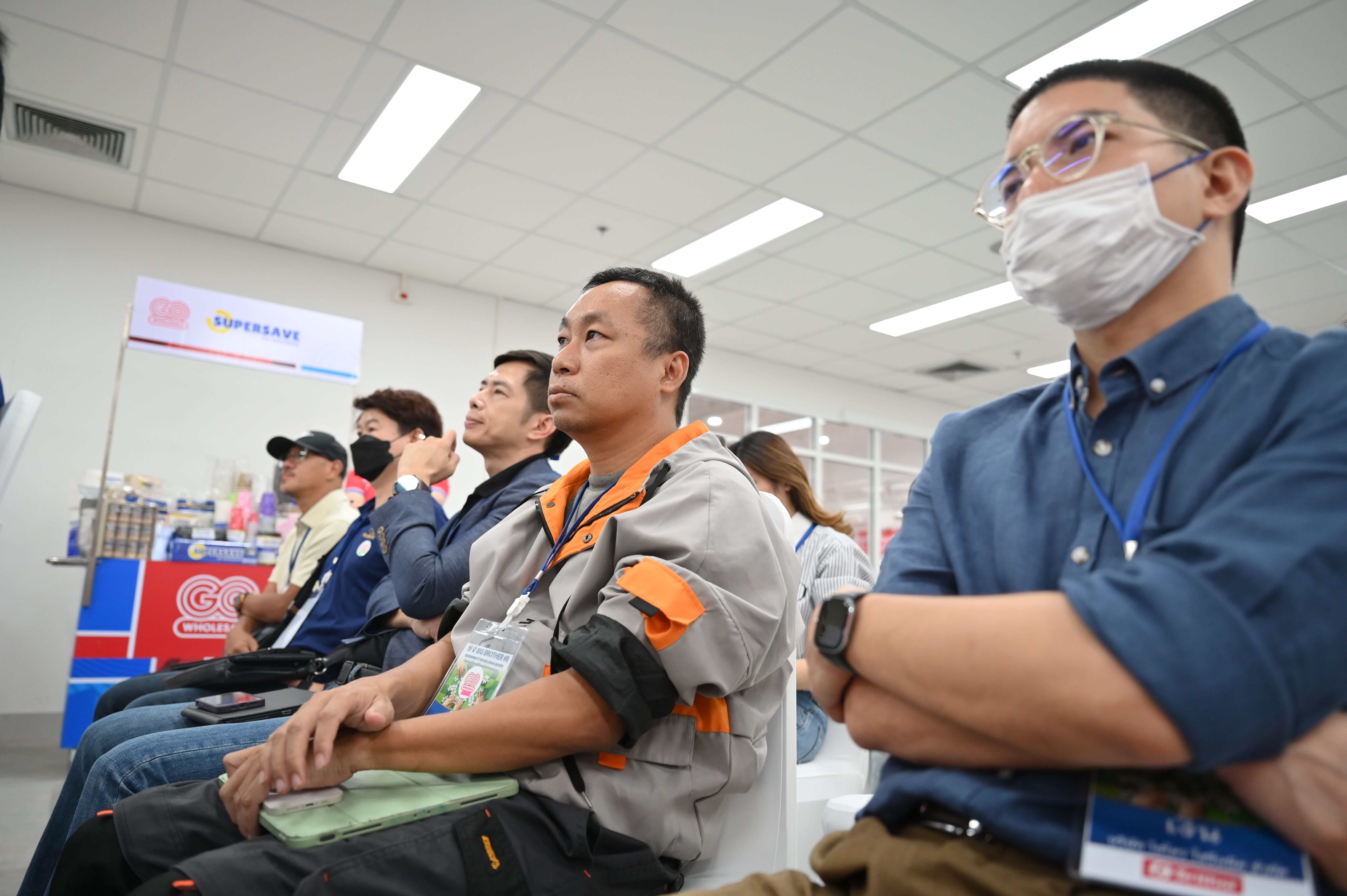 A diverse group of male attendees sits in a row, some wearing masks, observing a presentation.