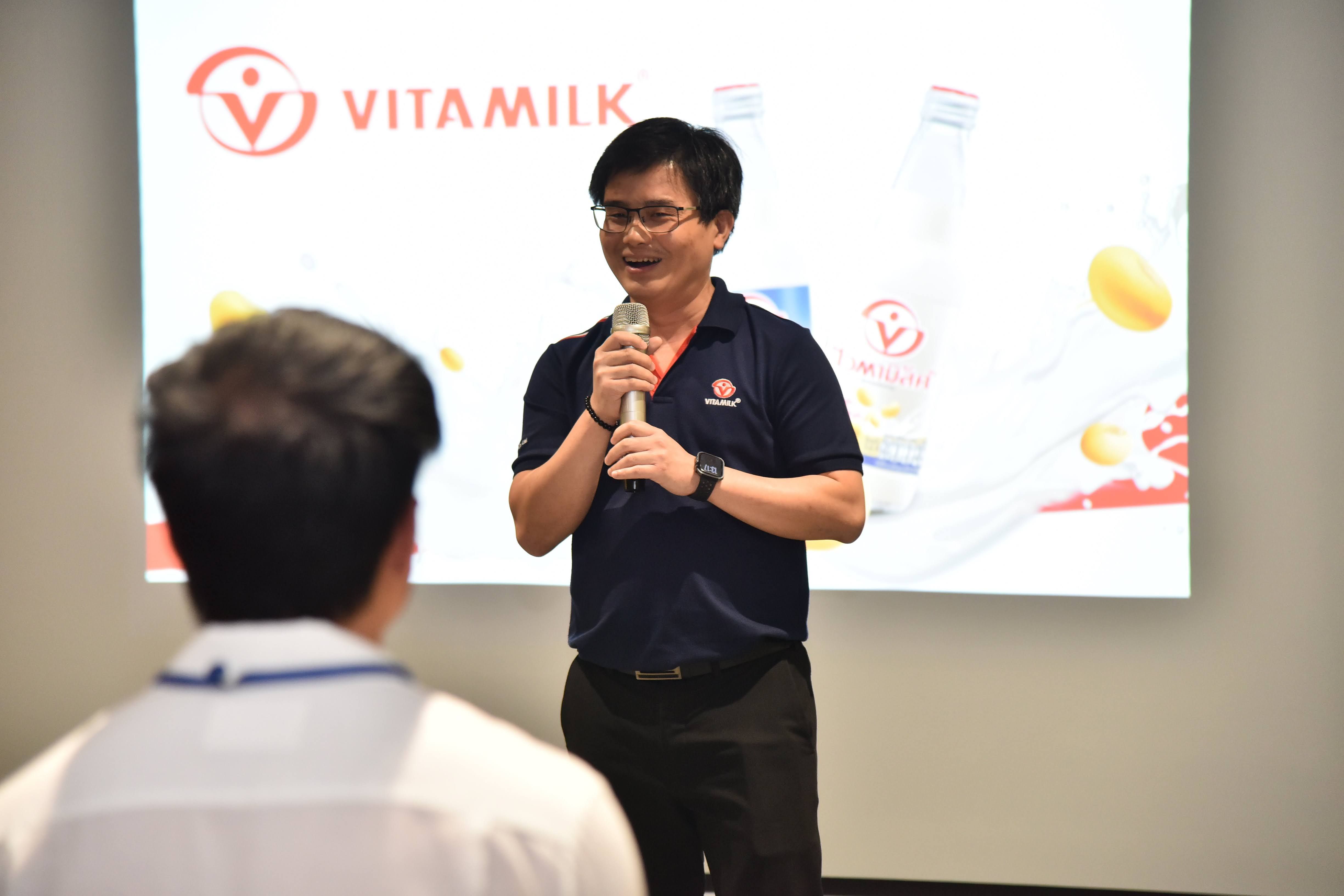 Man in navy polo shirt speaks into a microphone before a Vitamilk logo screen.