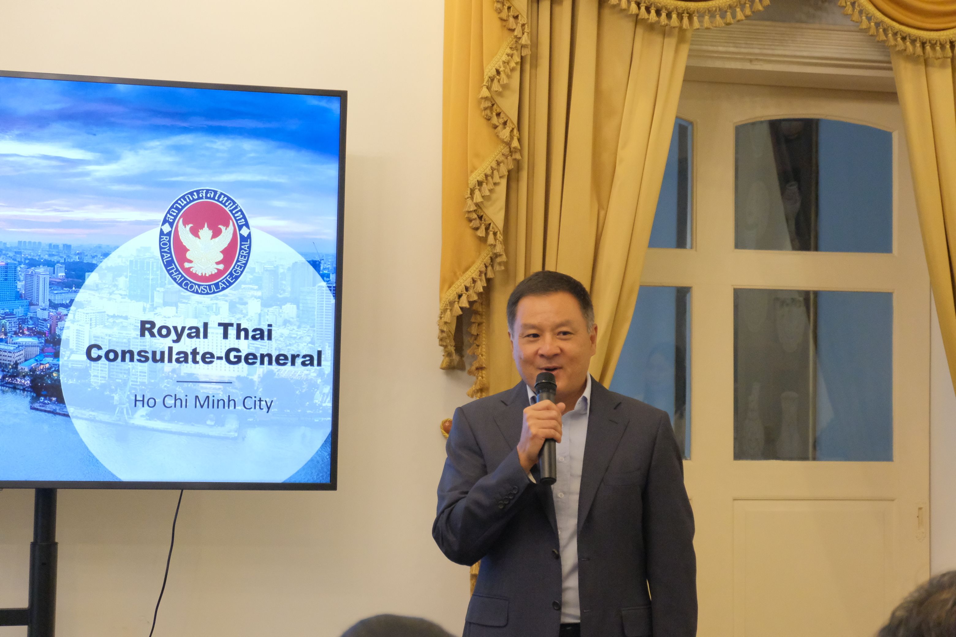 Man speaks into microphone in front of a screen displaying Royal Thai Consulate-General logo.