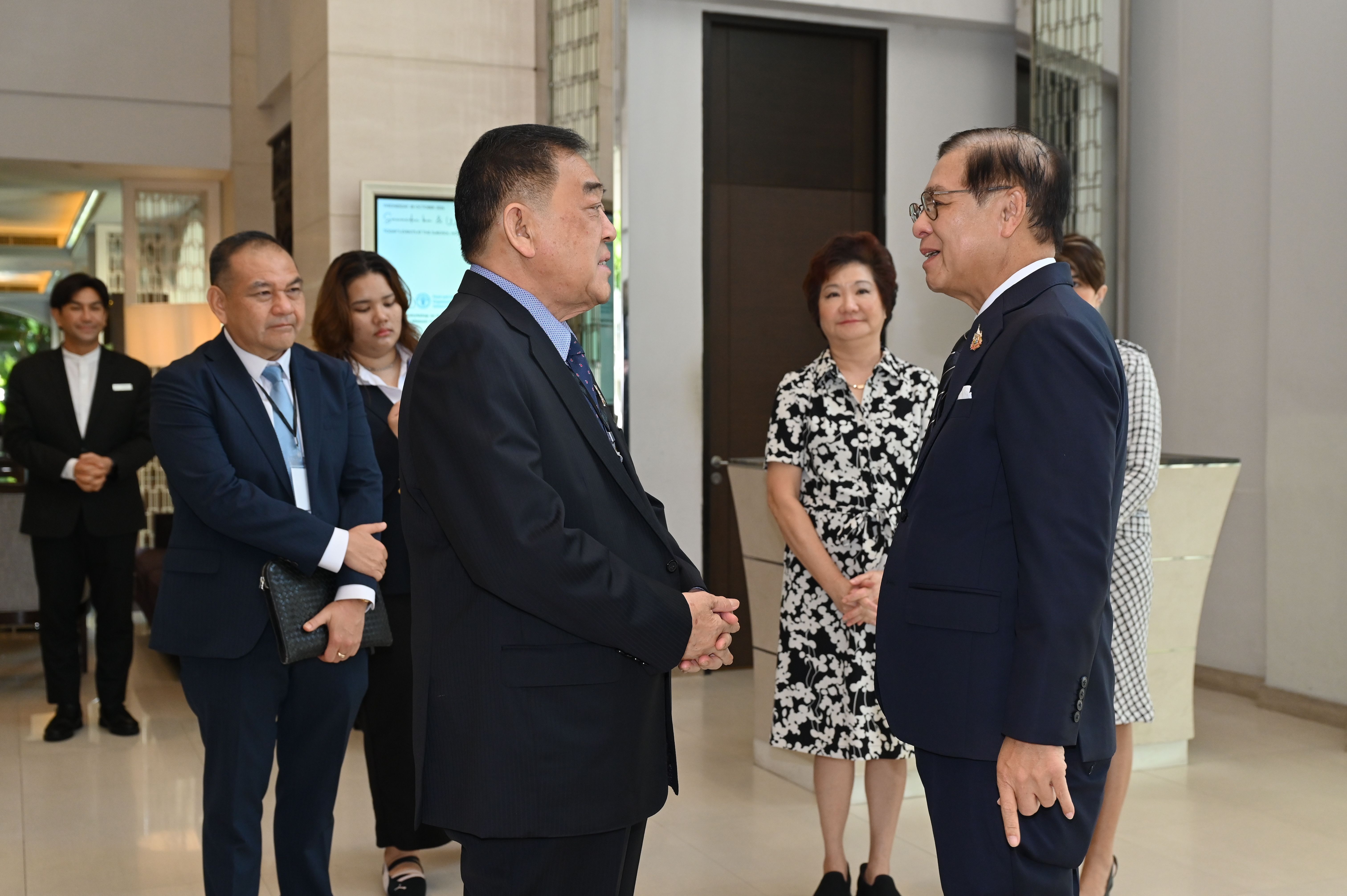 Two men in dark suits conversing in a modern indoor setting with other people.