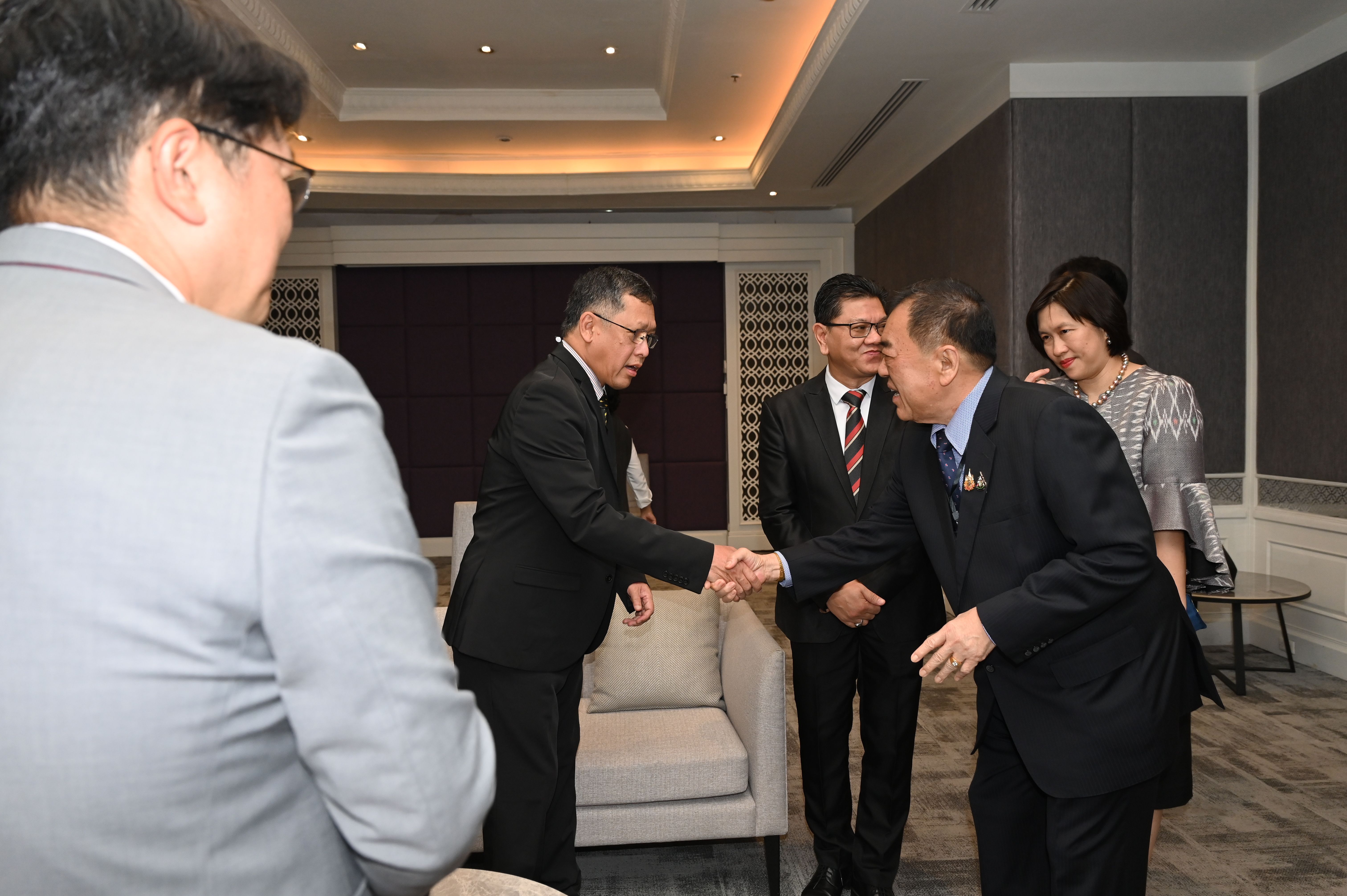 Two men in dark suits shake hands, one bowing slightly, at a formal indoor event.