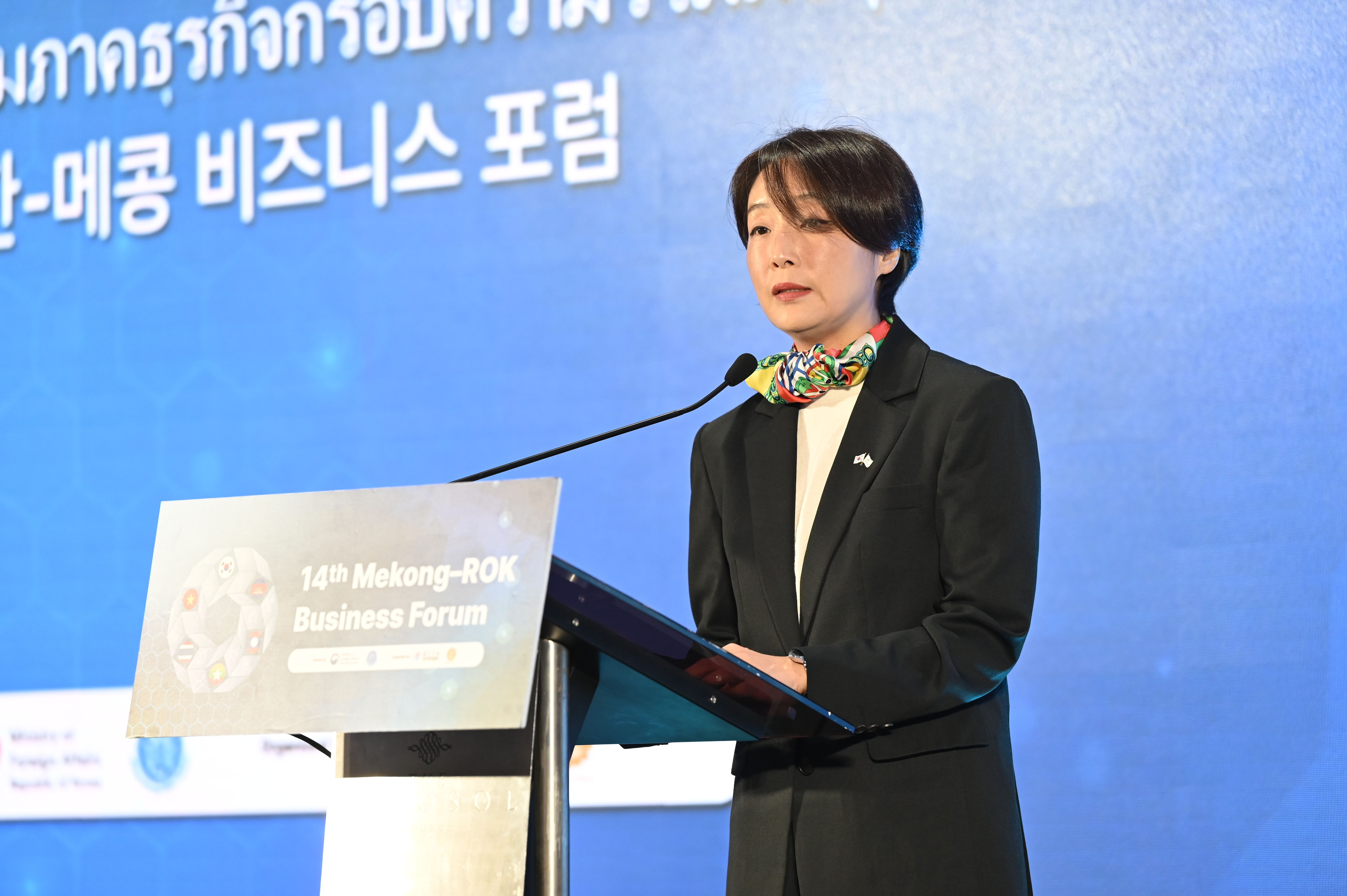 A woman in a dark suit speaks at a podium during an international business forum.