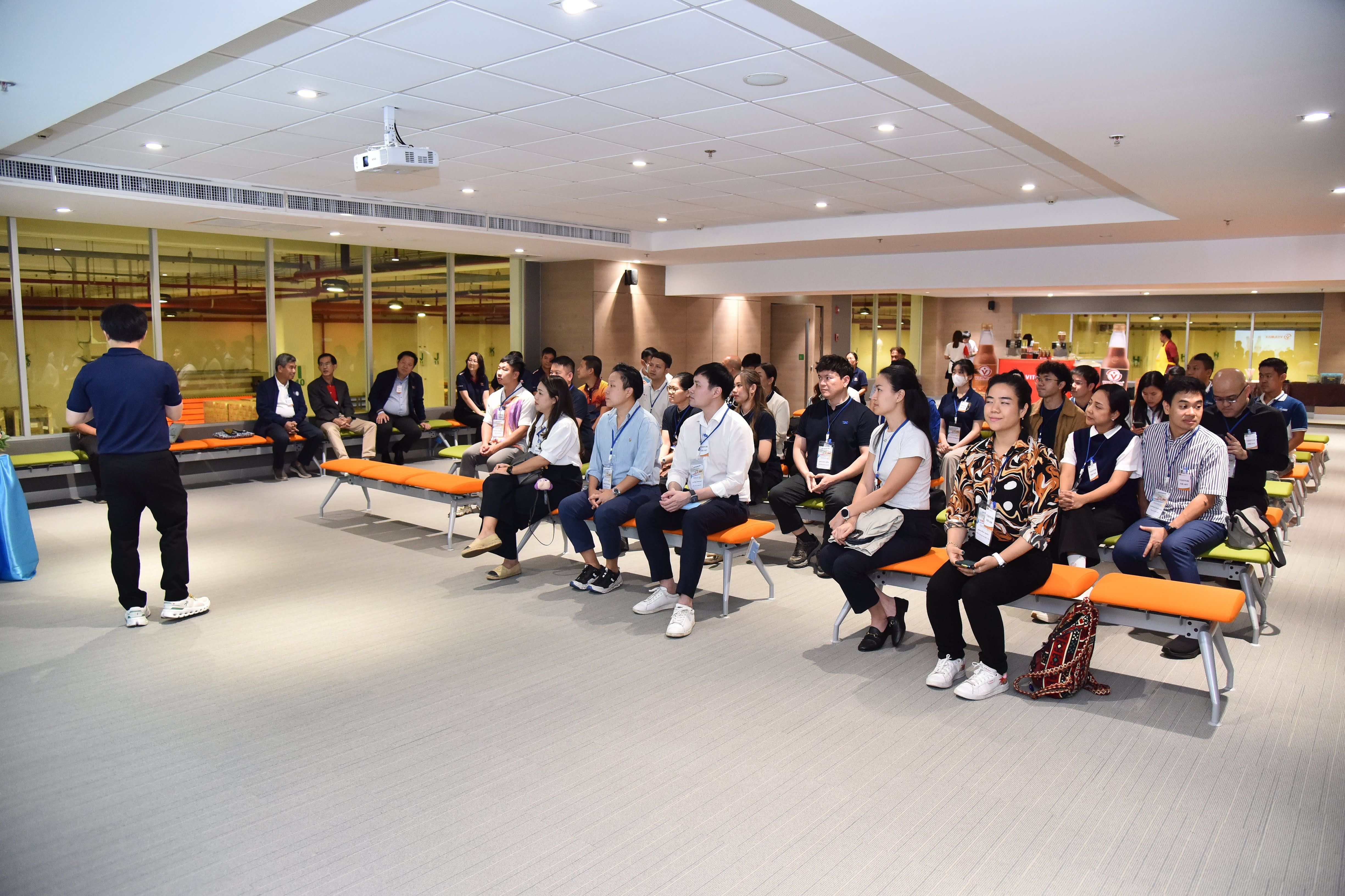 Speaker presents to an attentive audience seated on benches in a modern conference room.