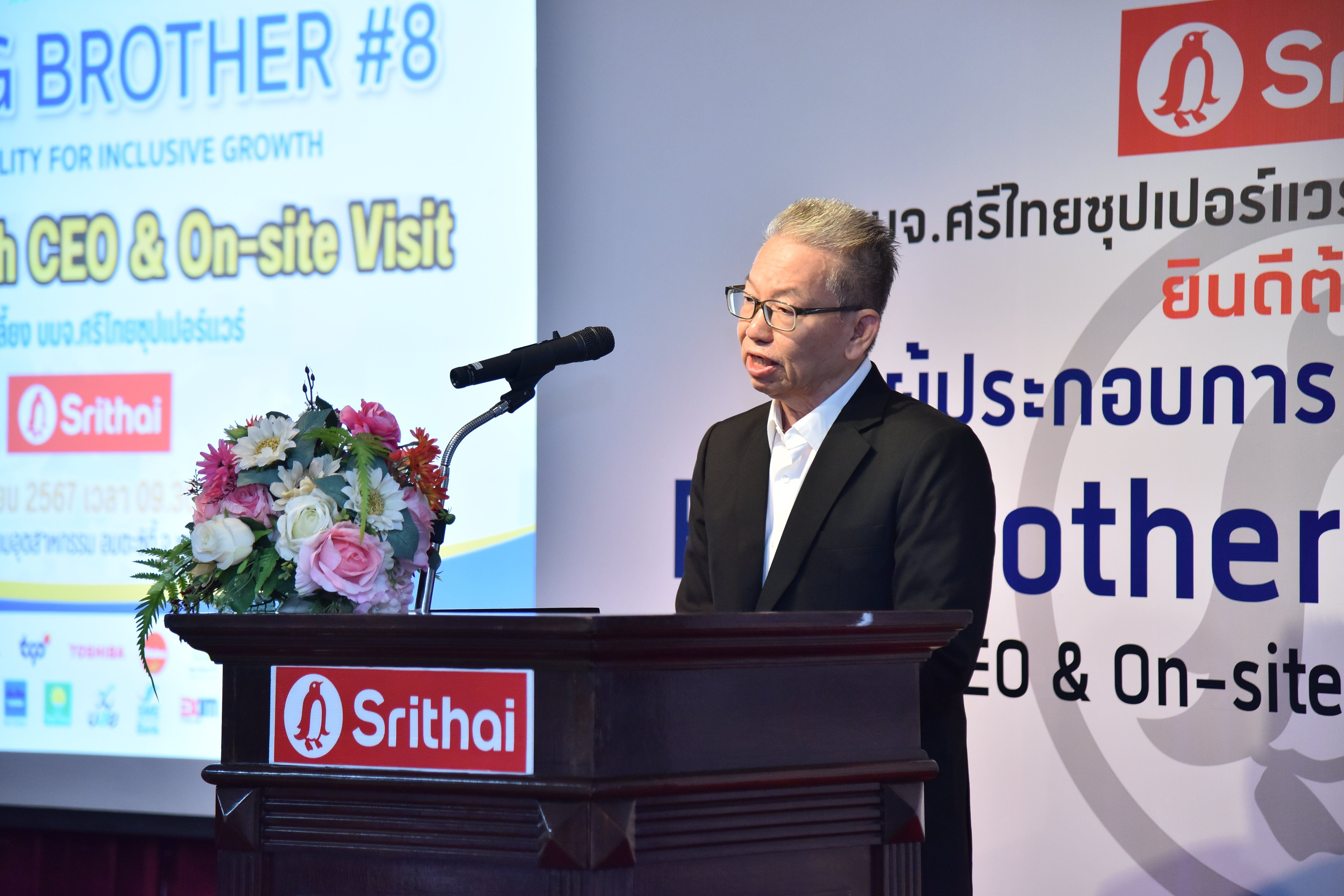 Man in suit and glasses speaks at a podium during a Srithai event presentation.