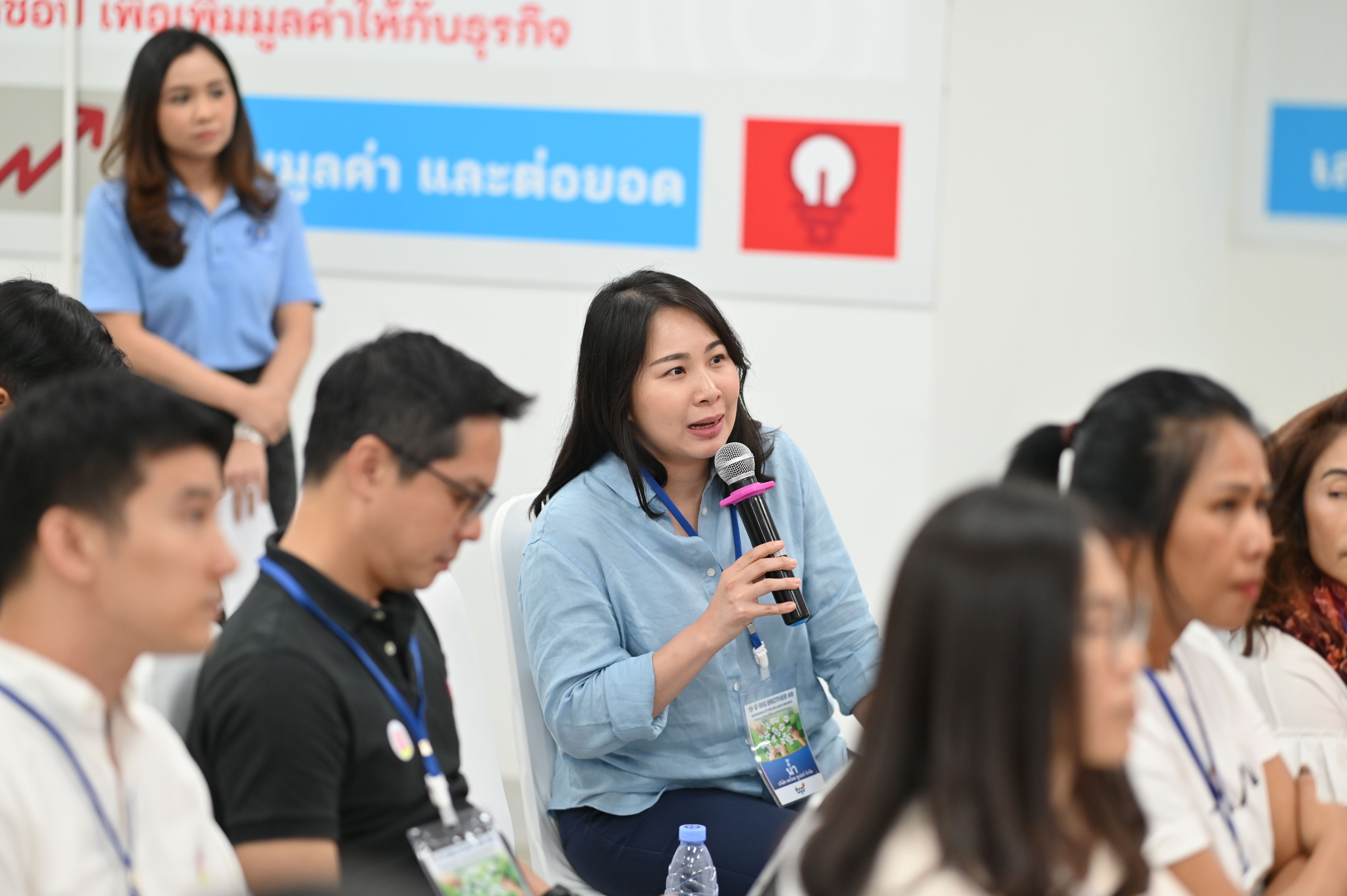 A woman speaks into a microphone at a seminar, surrounded by attendees.