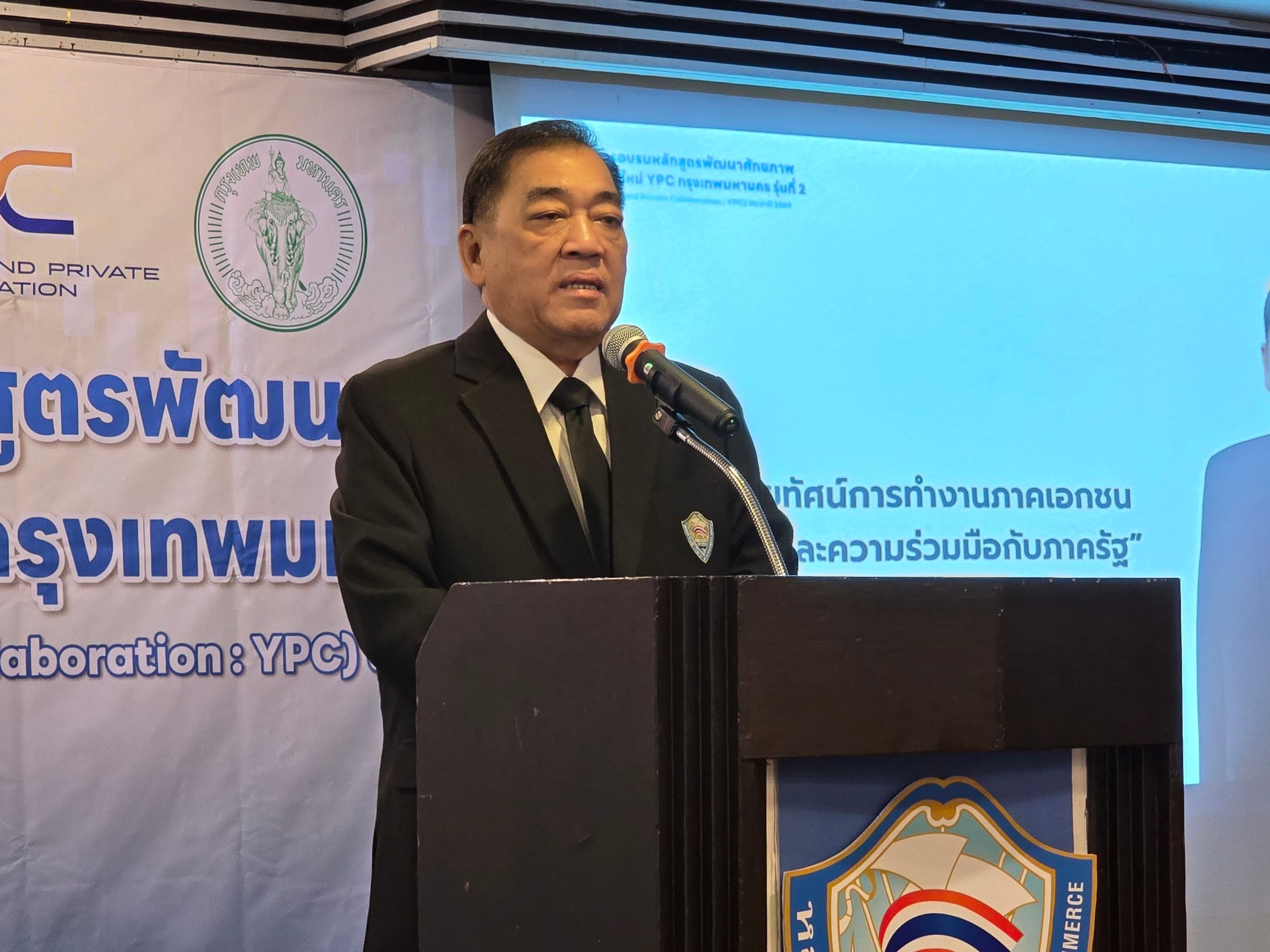 A man in a dark suit speaks into a microphone at a podium during a presentation.