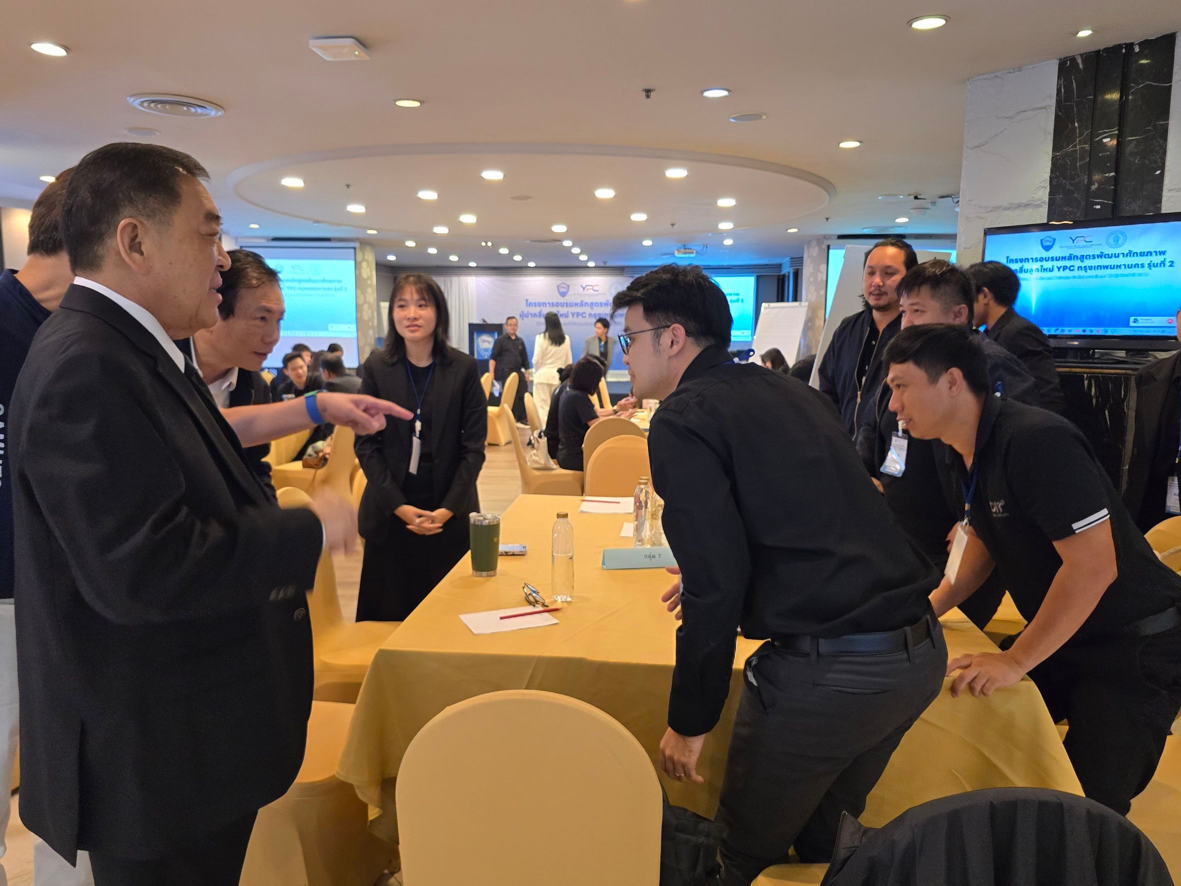 An older man speaks to a group of professionals in a busy conference room.
