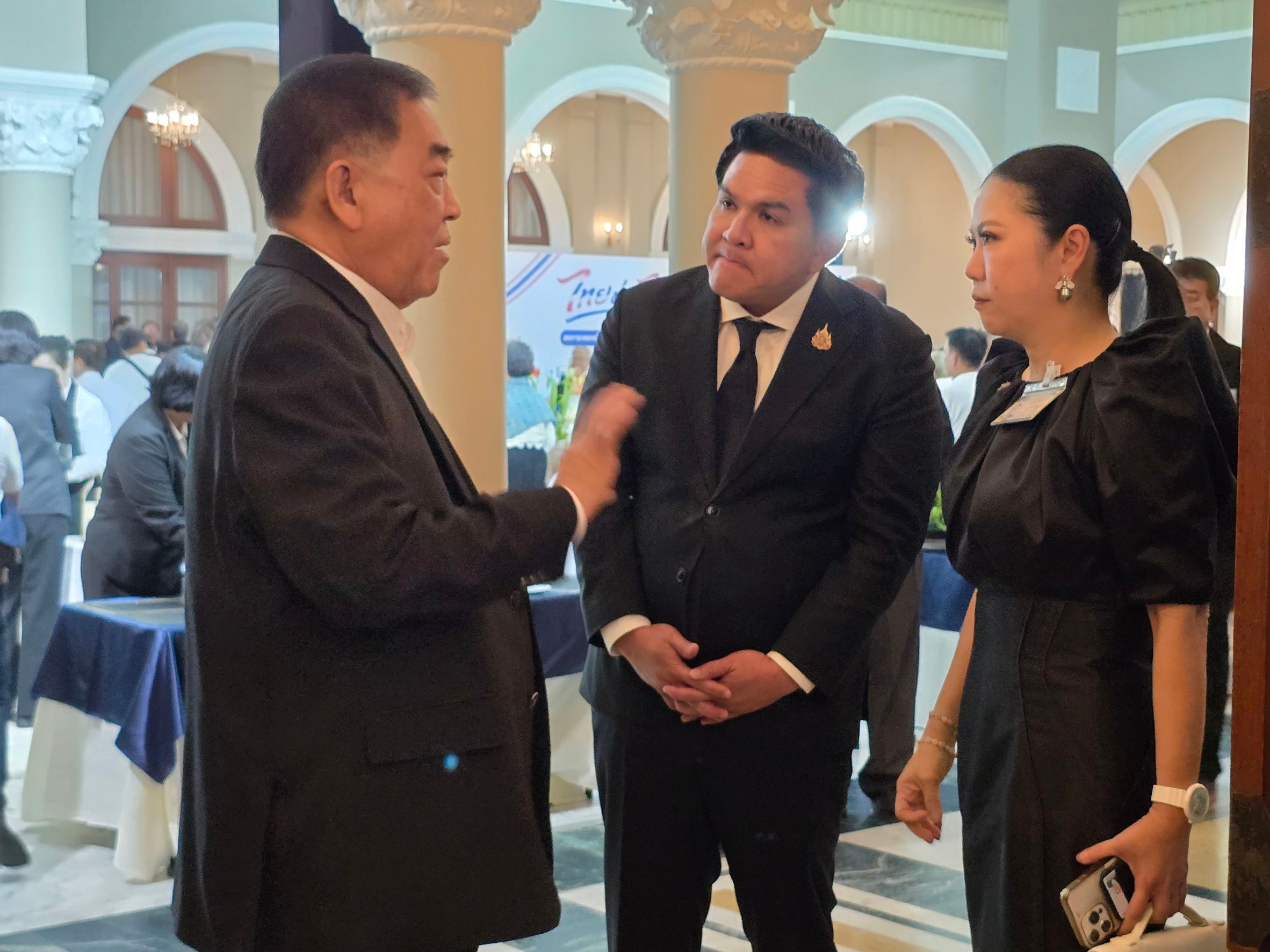 Three elegantly dressed individuals, two men and one woman, conversing at an indoor event.