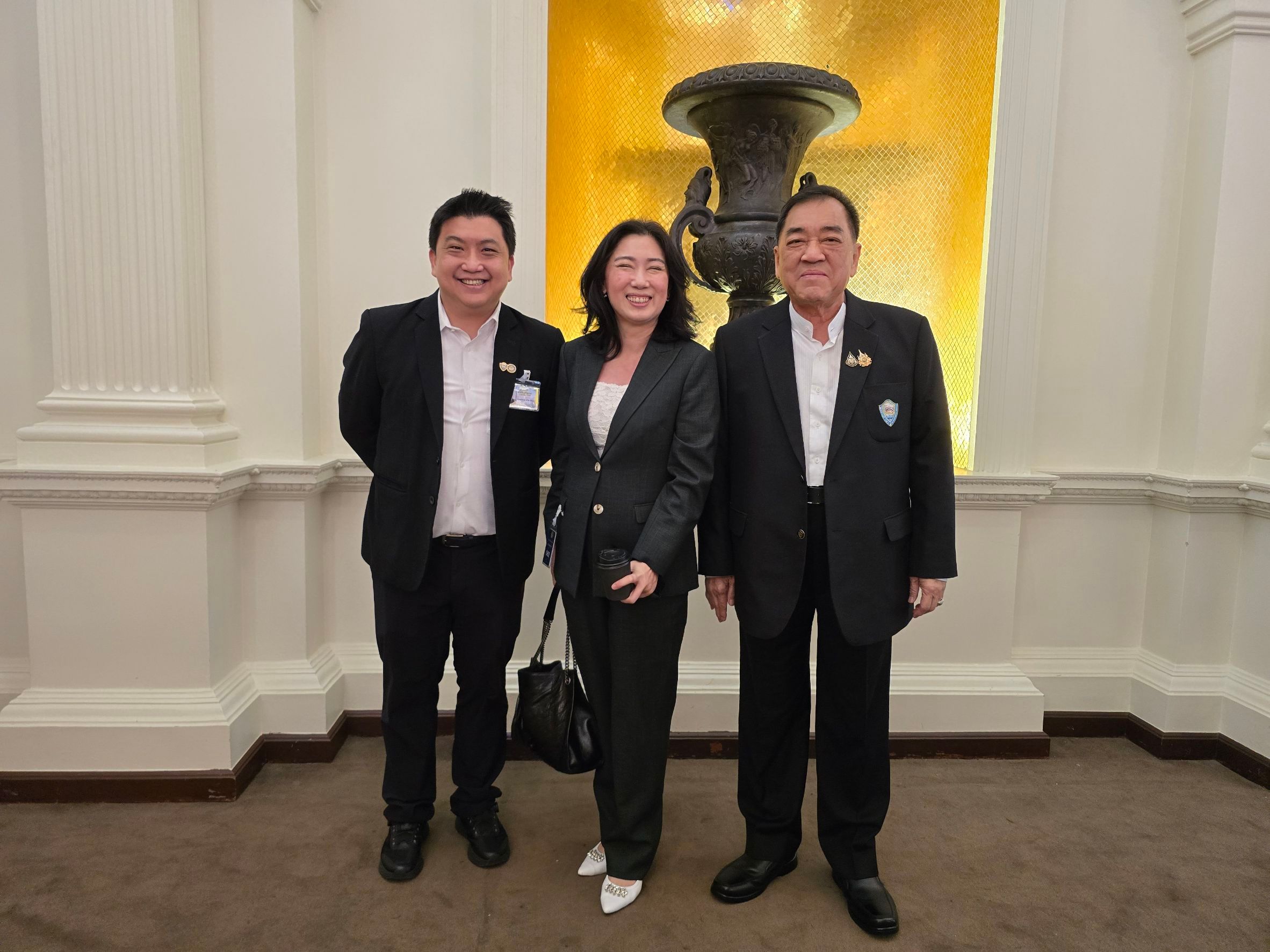 Three smiling professionals, two men and one woman, stand formally against a golden decorative wall.