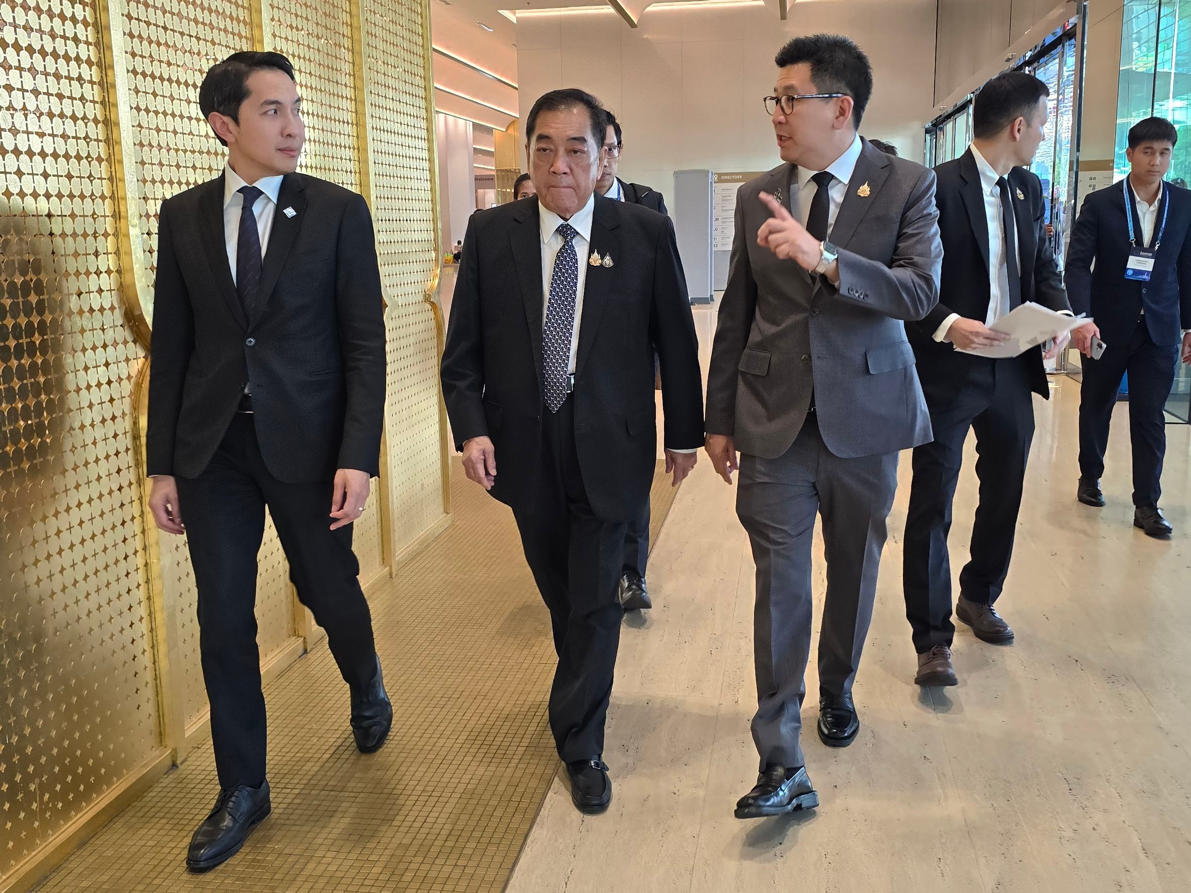 Three men in dark suits walk through a modern building with gold textured walls.
