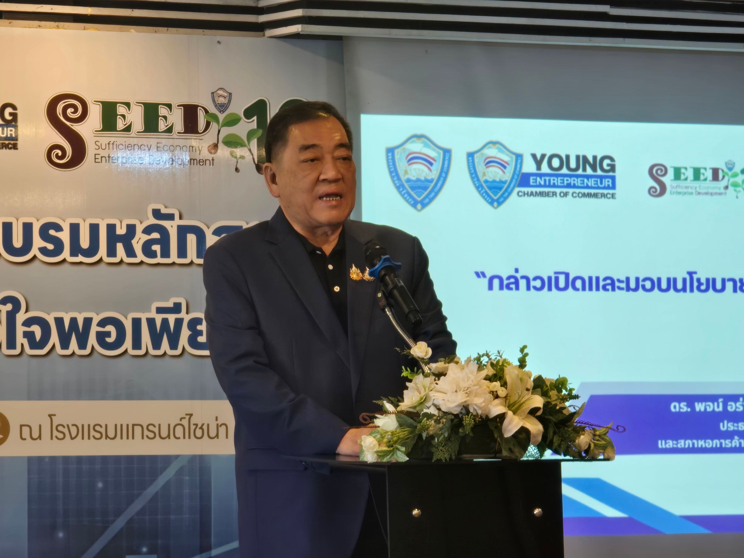 Man in a dark suit speaks at a podium with event logos and Thai text in the background.