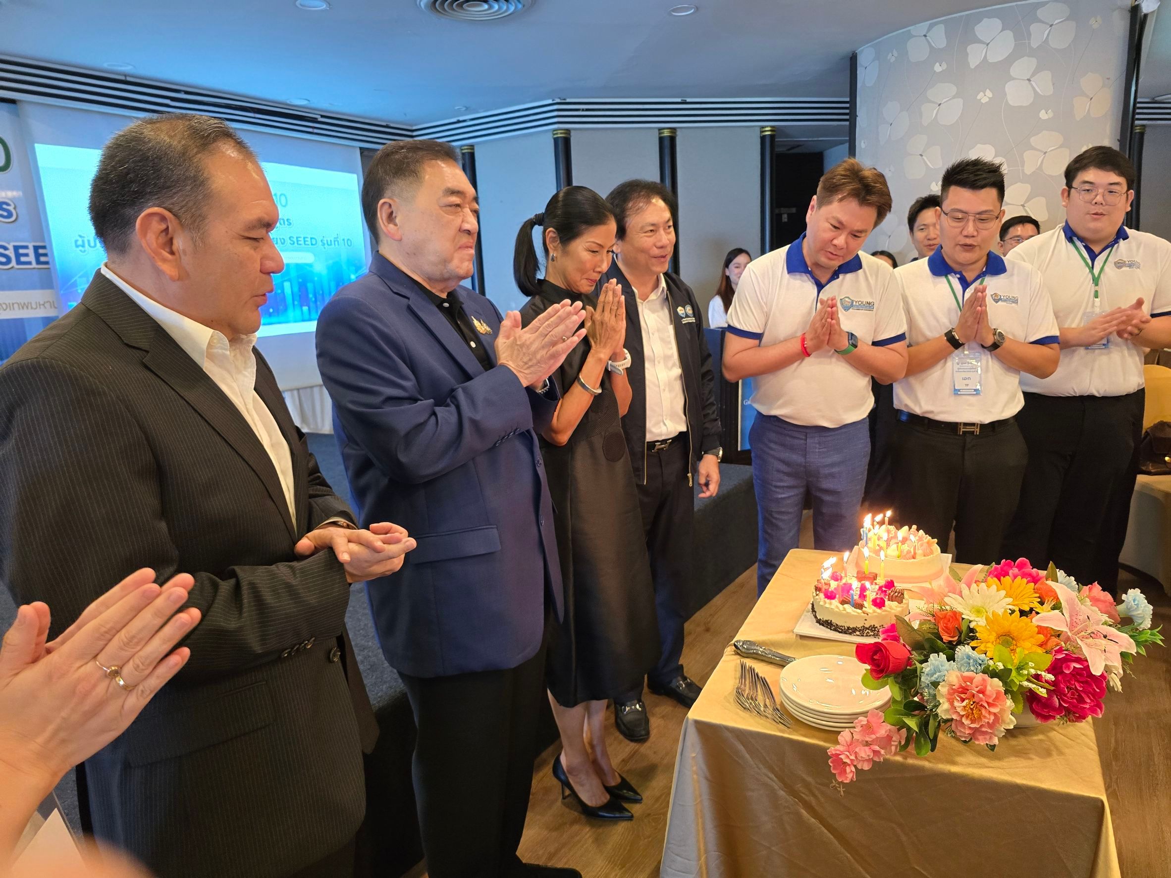 Group of people celebrating around a birthday cake with some clapping and others wai gesturing.
