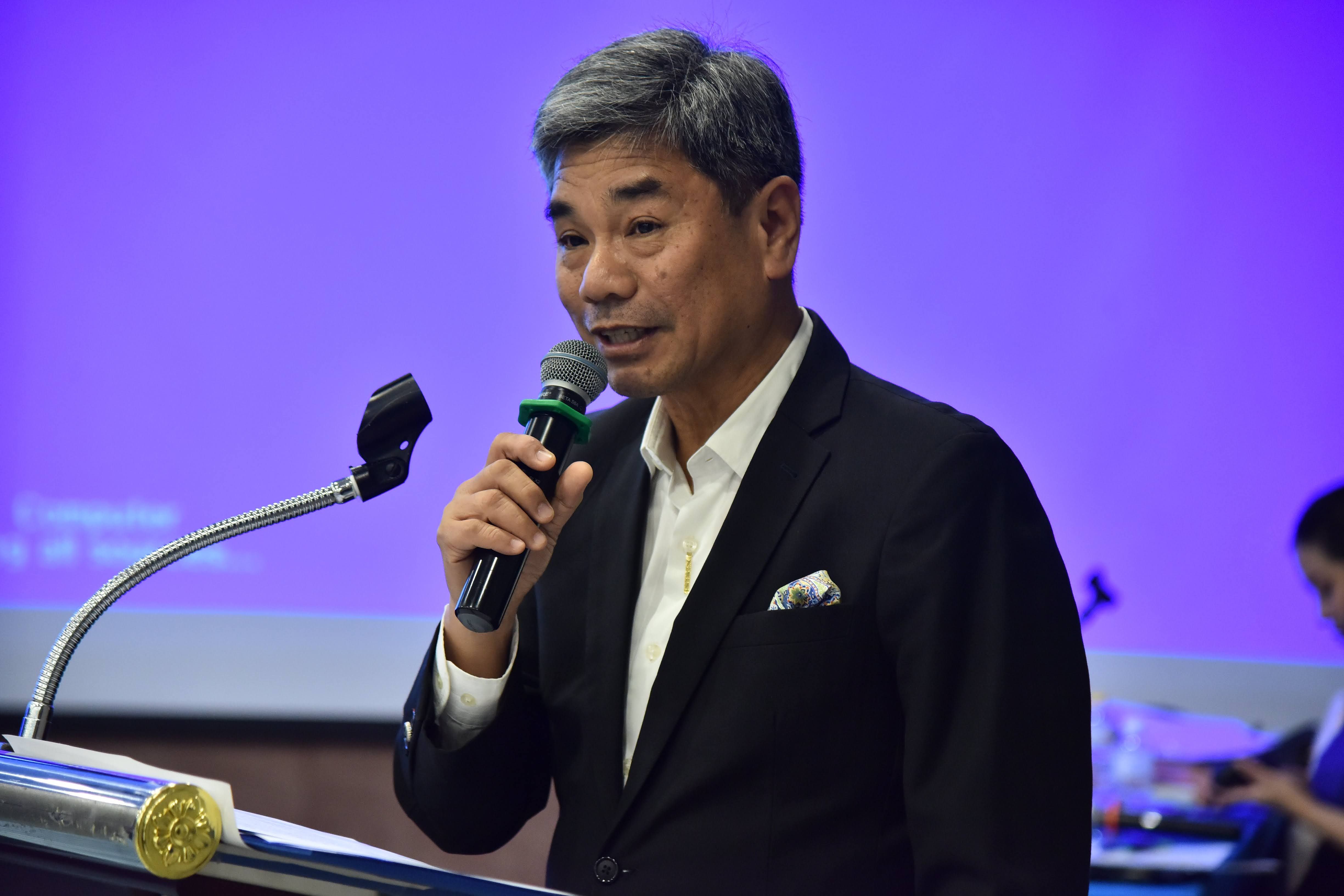 Asian man with grey hair speaking into a microphone at a podium.