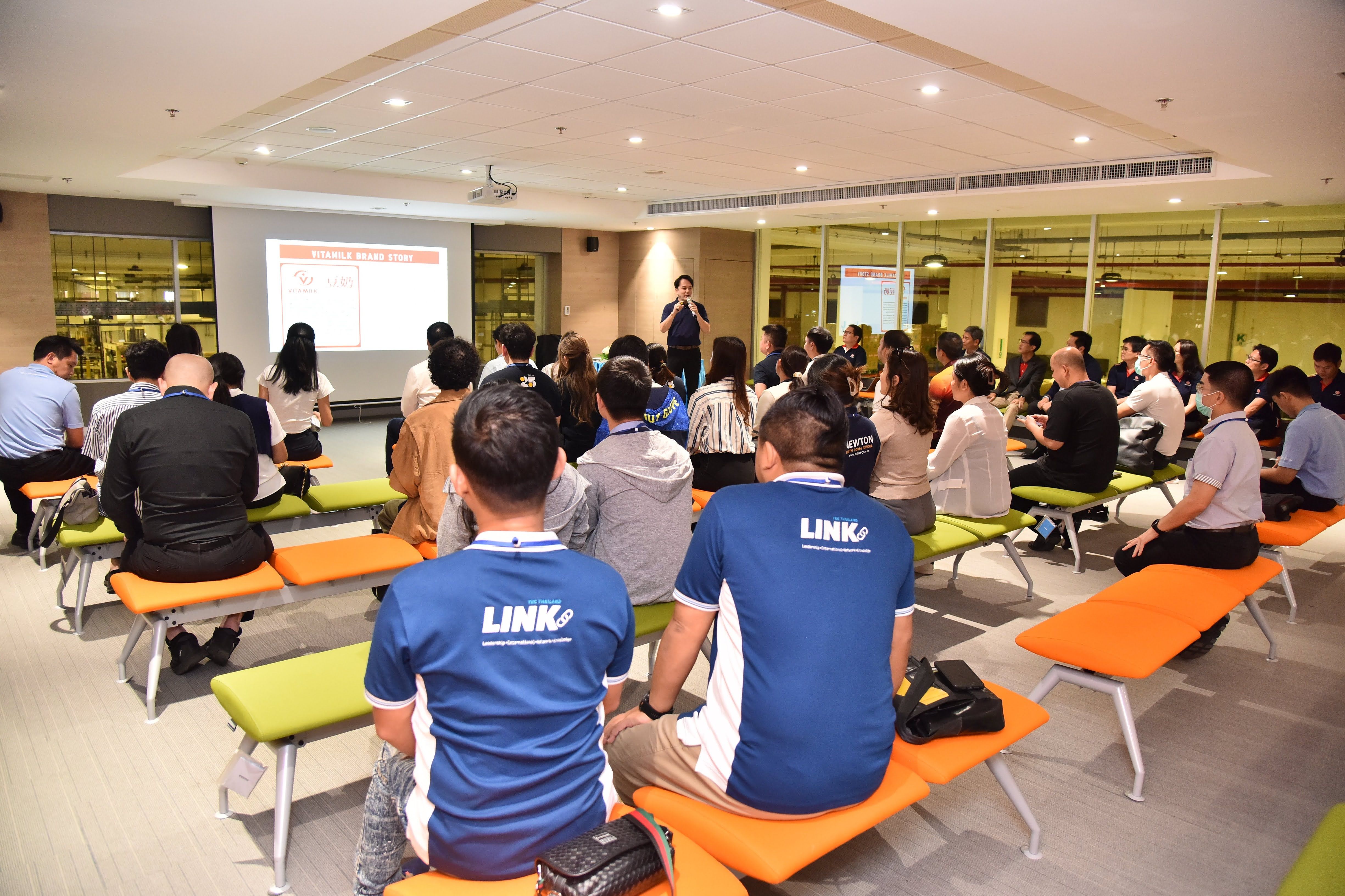 Audience members watch a presenter and screen in a modern conference room during a seminar.
