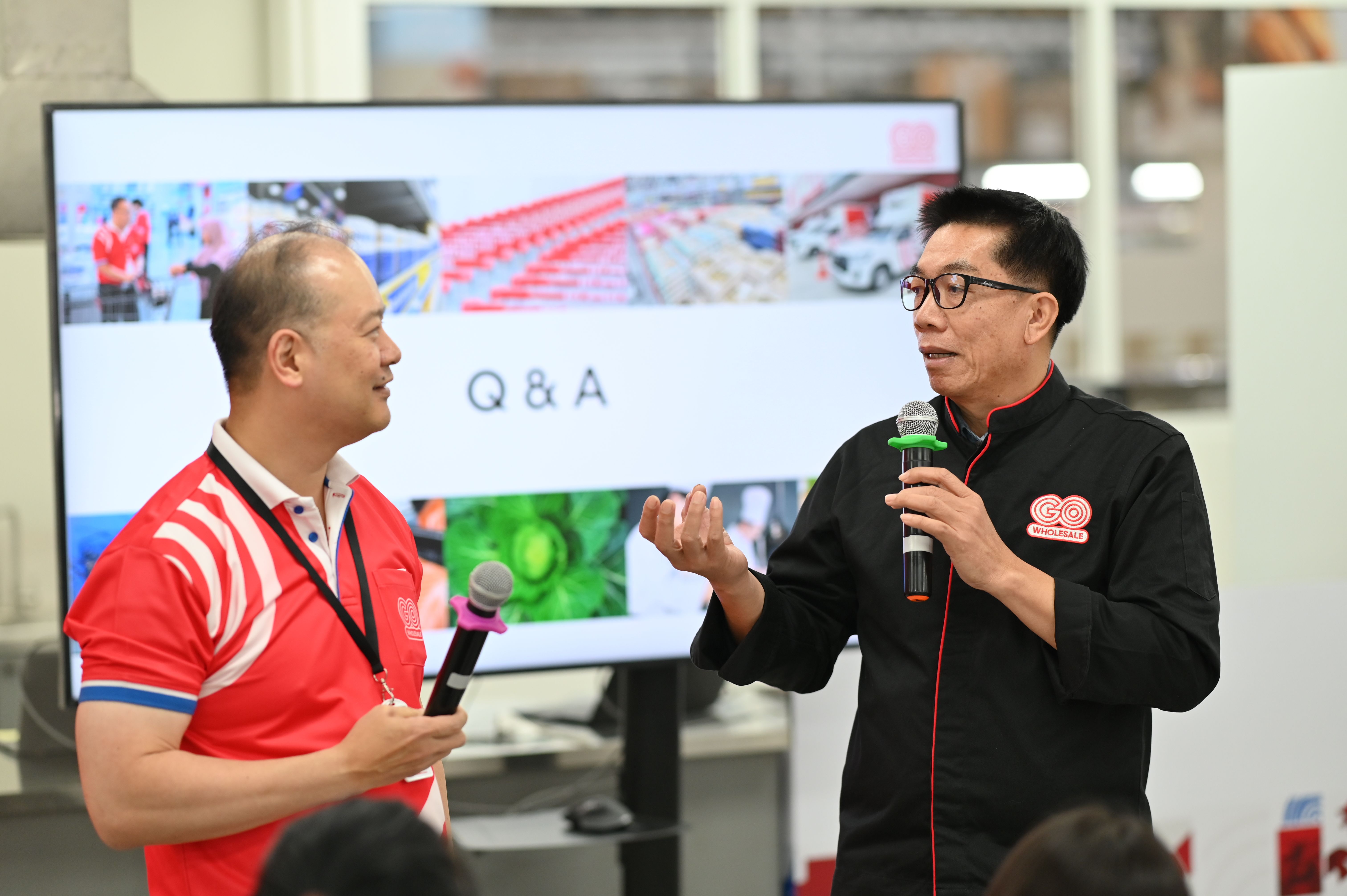 Two men hold microphones, discussing during a Q&A session with a large screen.