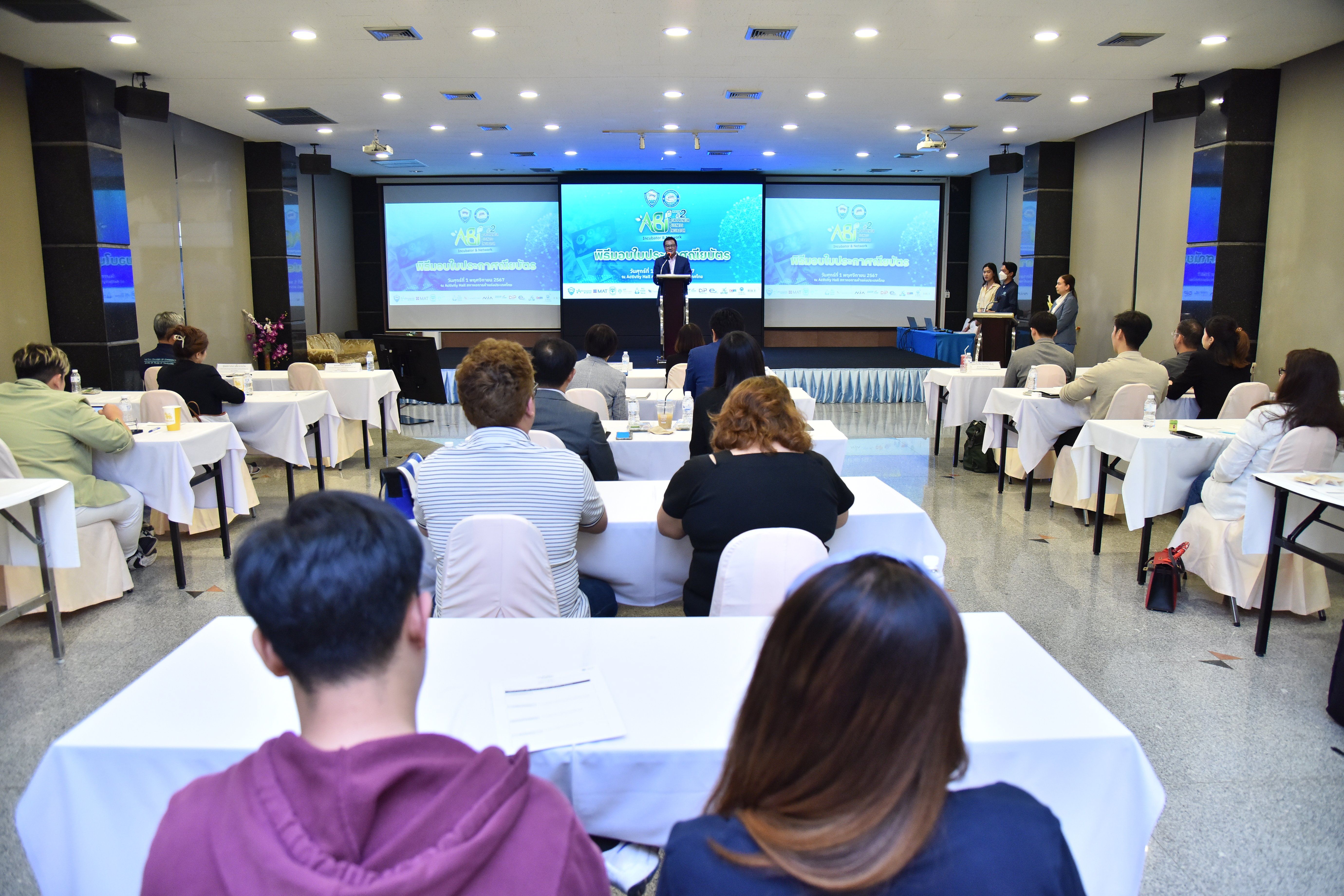 A speaker presents to an audience in a conference room with large screens.