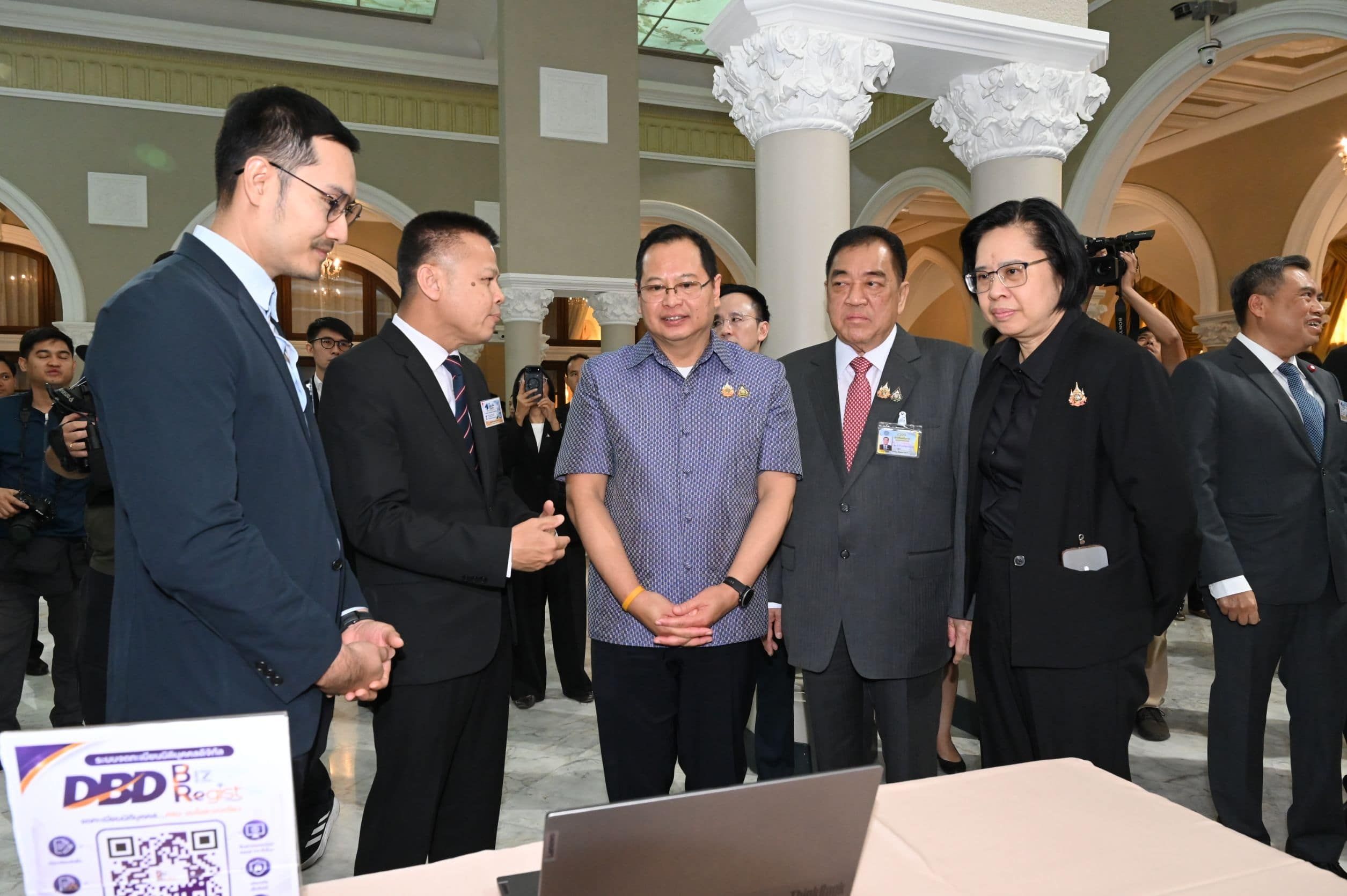 Officials gather around a laptop presentation at an indoor event with pillars.