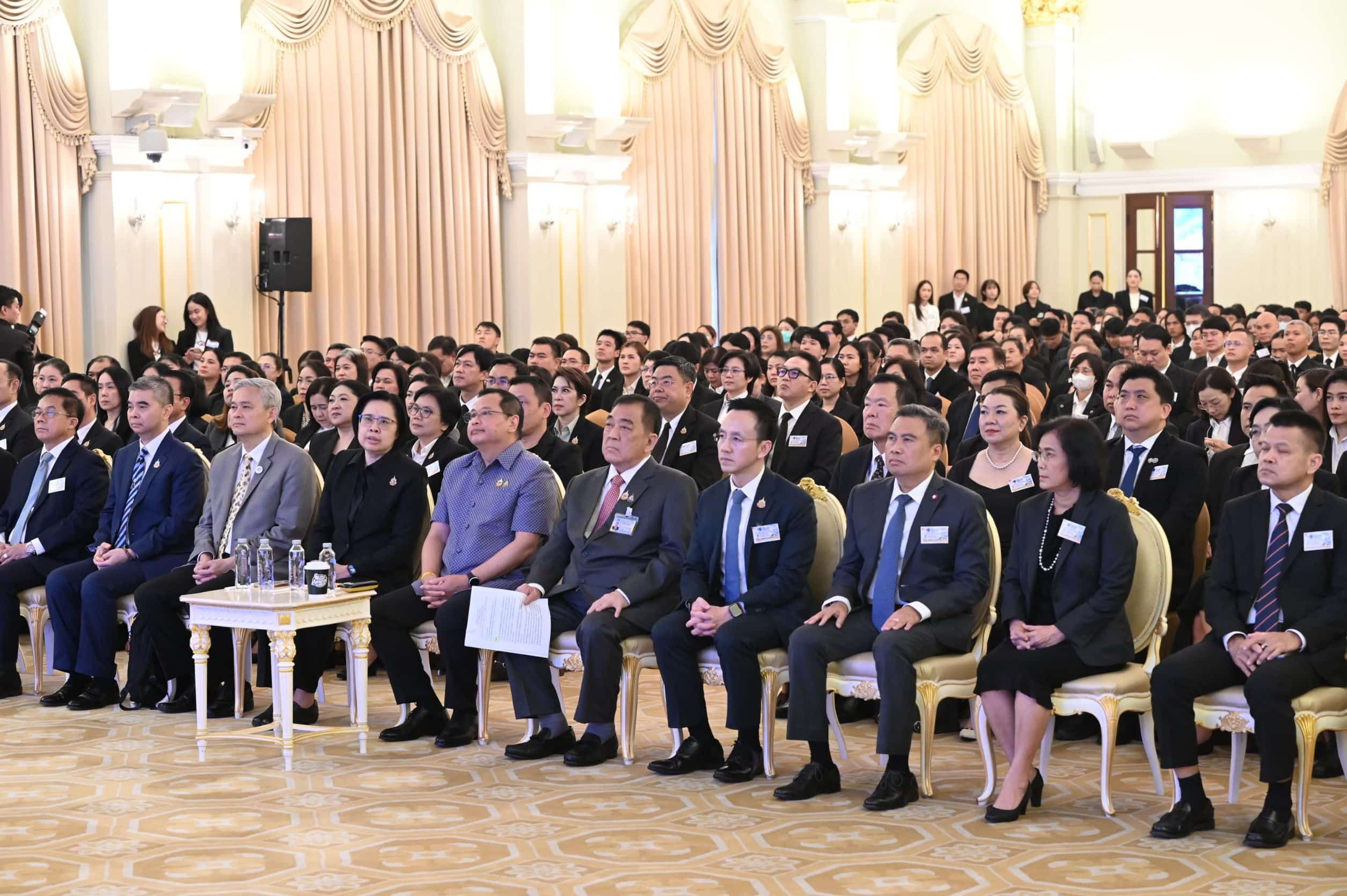 A large audience of formally dressed people sits attentively in a grand hall.