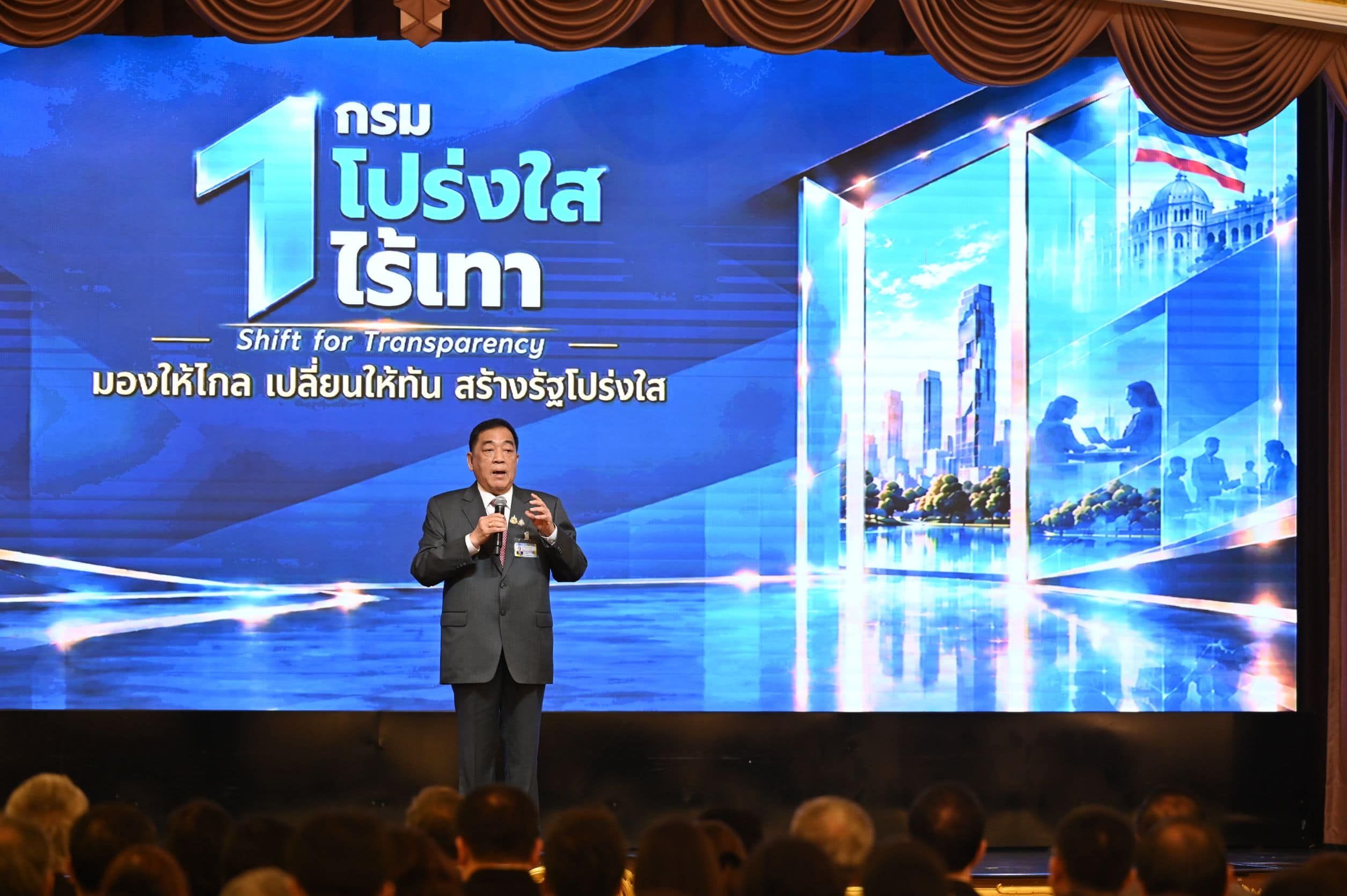 Man in a suit speaks on stage in front of a screen displaying 'Shift For Transparency'.