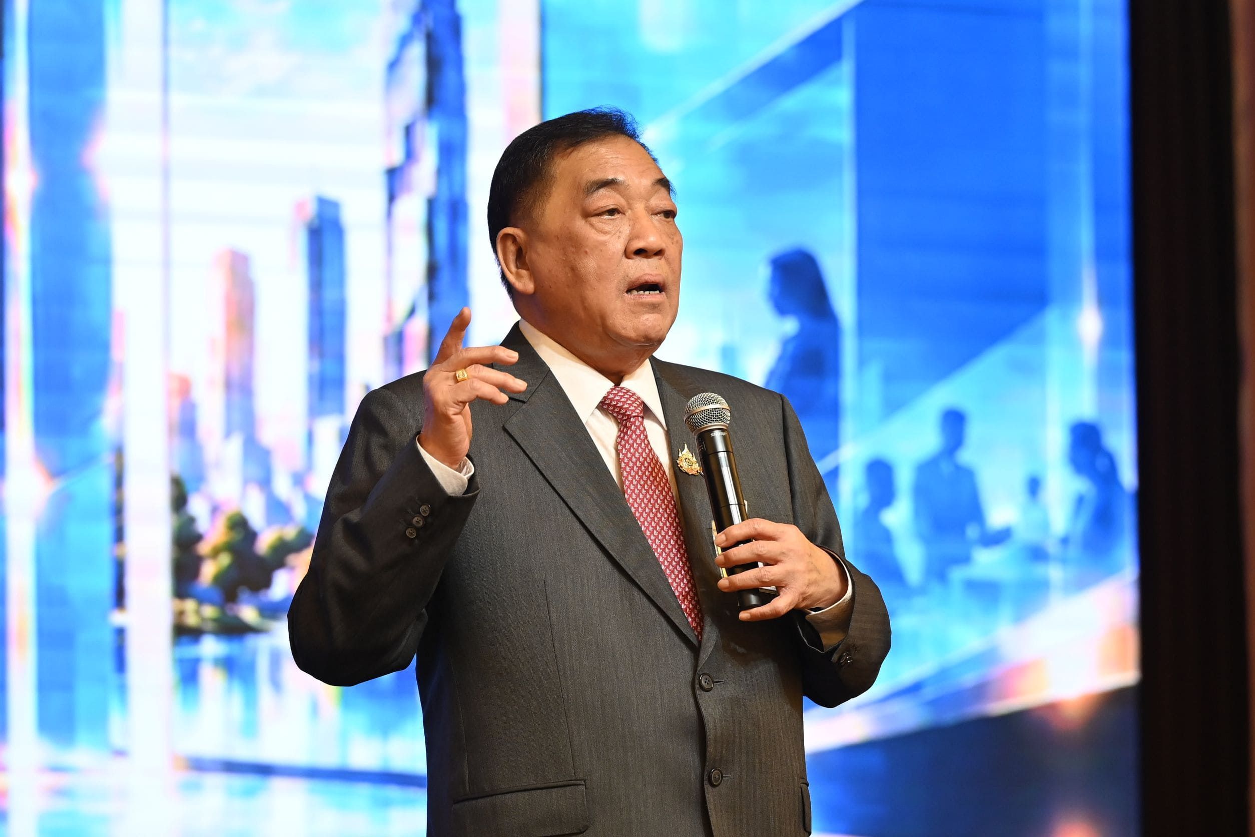 A man in a dark suit speaks into a microphone, gesturing during a presentation.