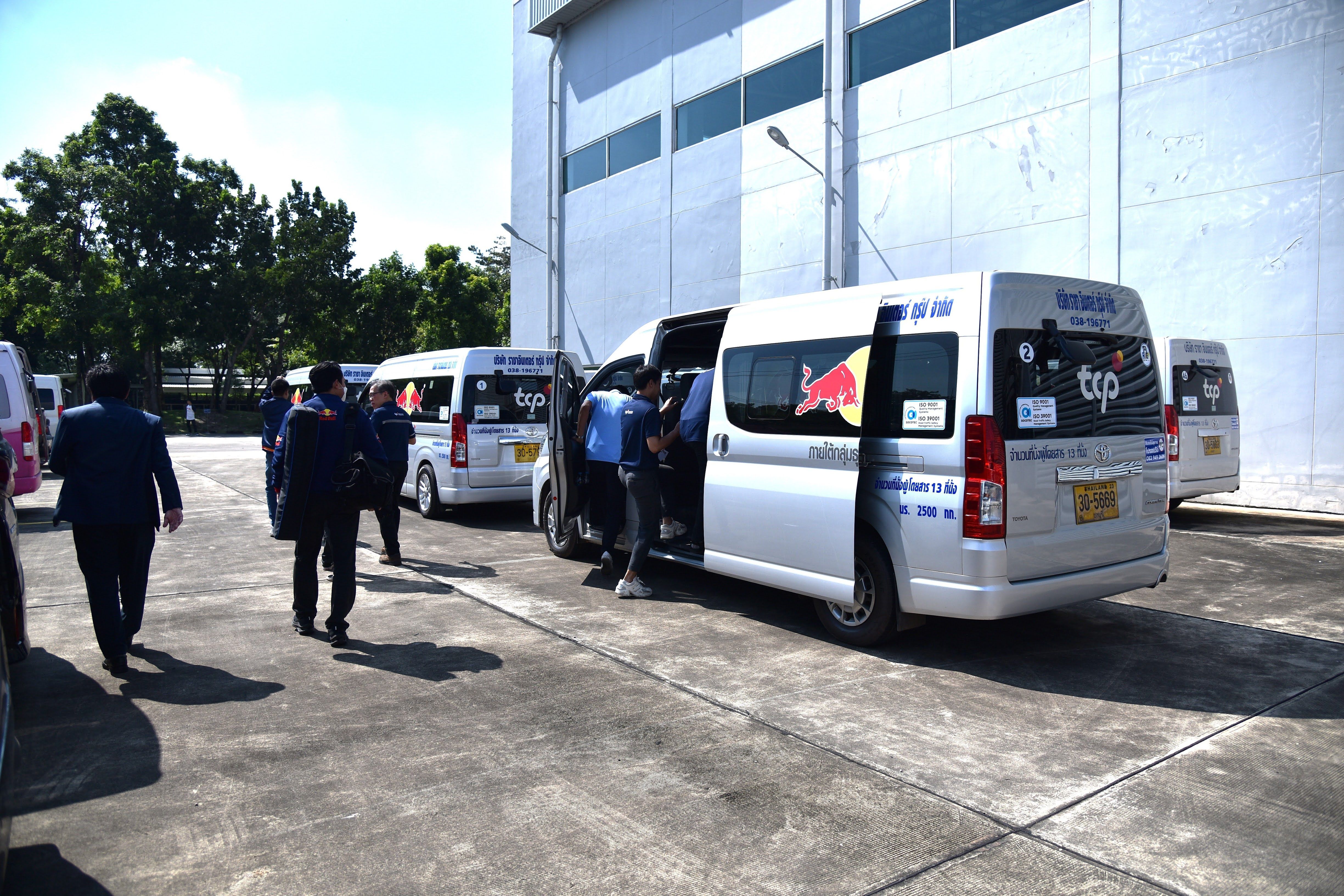 People board a silver van with its side door open, parked alongside other white vans.