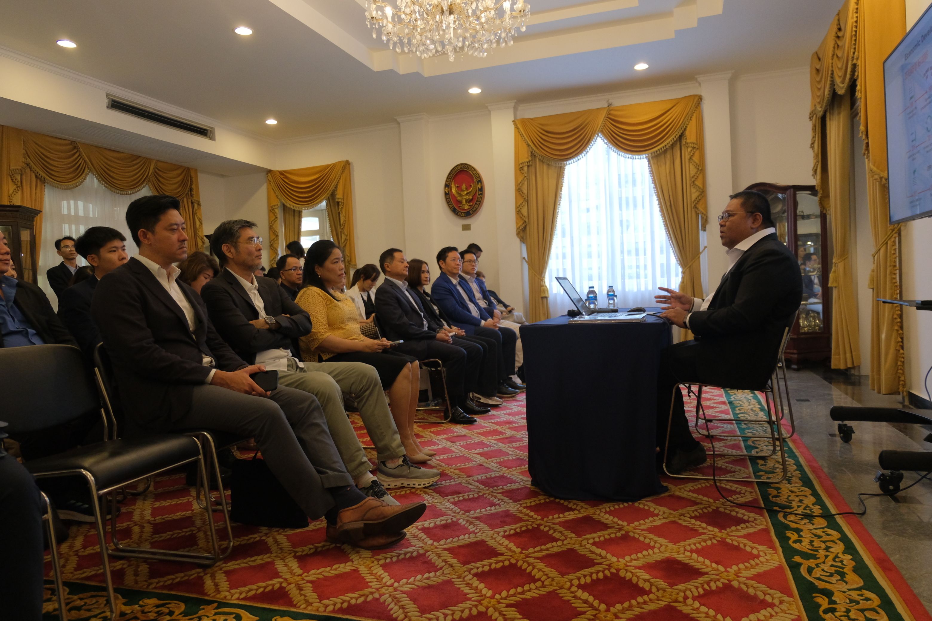 Man speaking to a seated audience in a formal conference room with a chandelier.