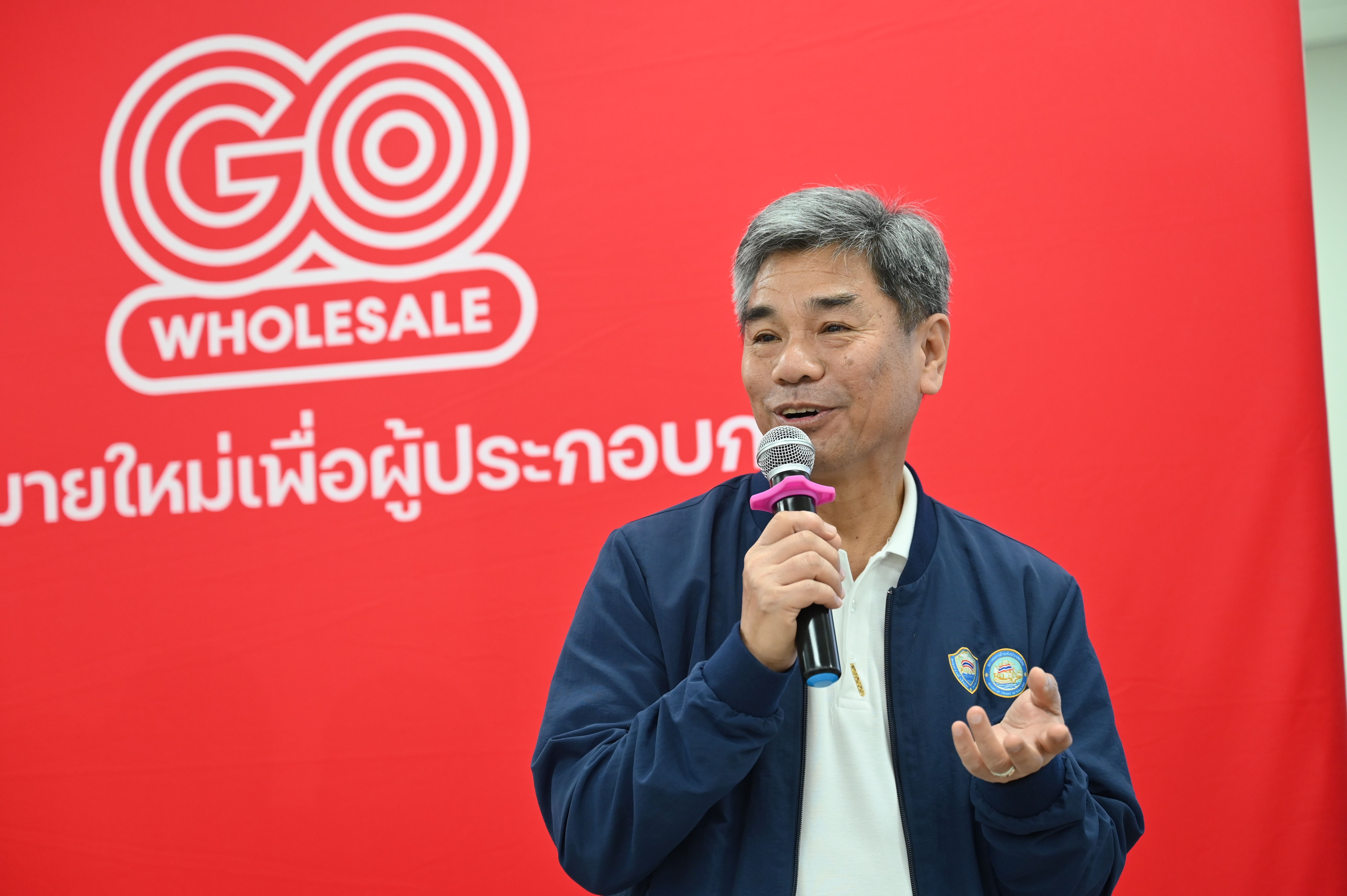 Man in blue jacket speaks into microphone in front of a red 'GO WHOLESALE' banner.