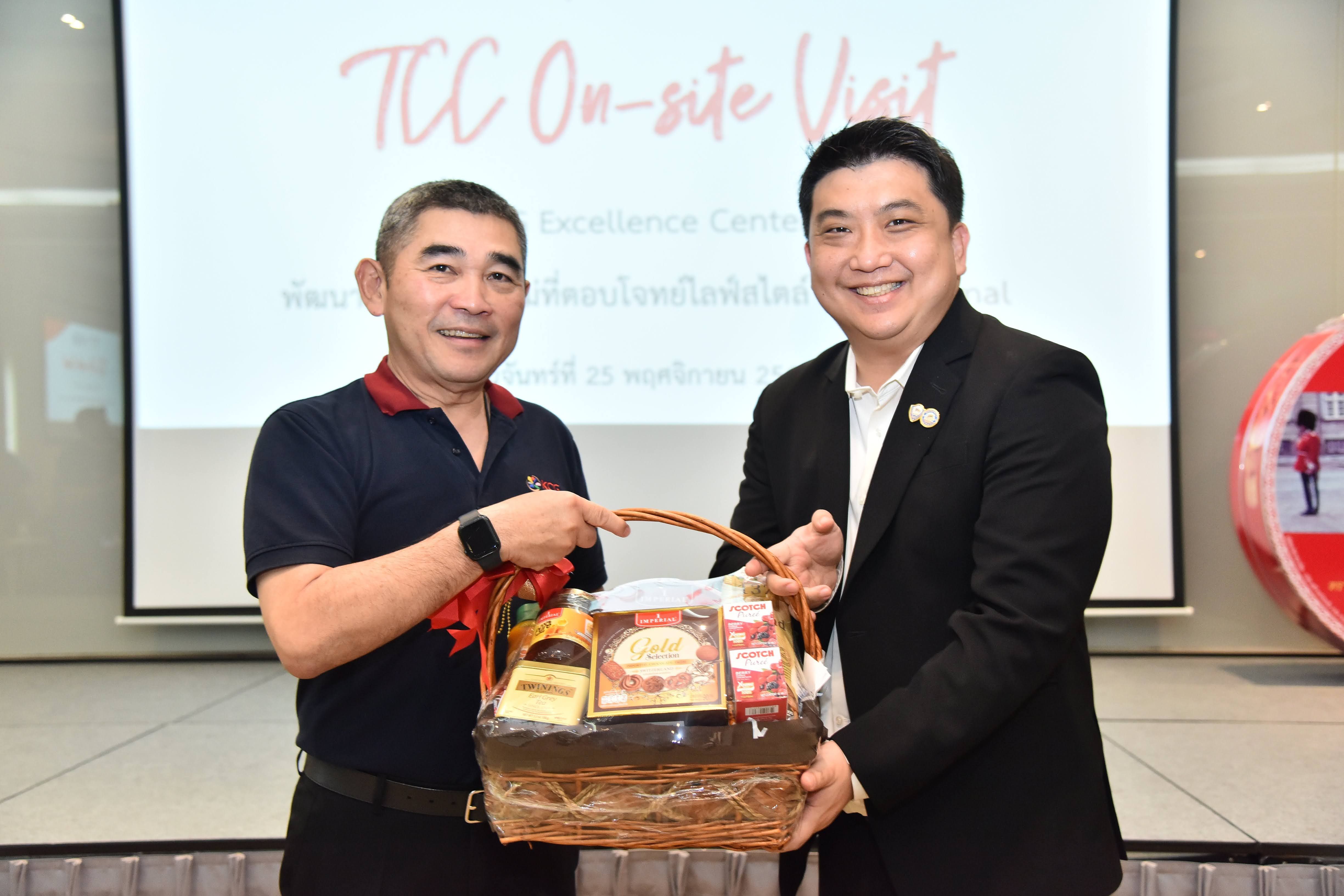Two men exchange a gift basket with "TCC On-site Visit" text visible on a screen in the background.