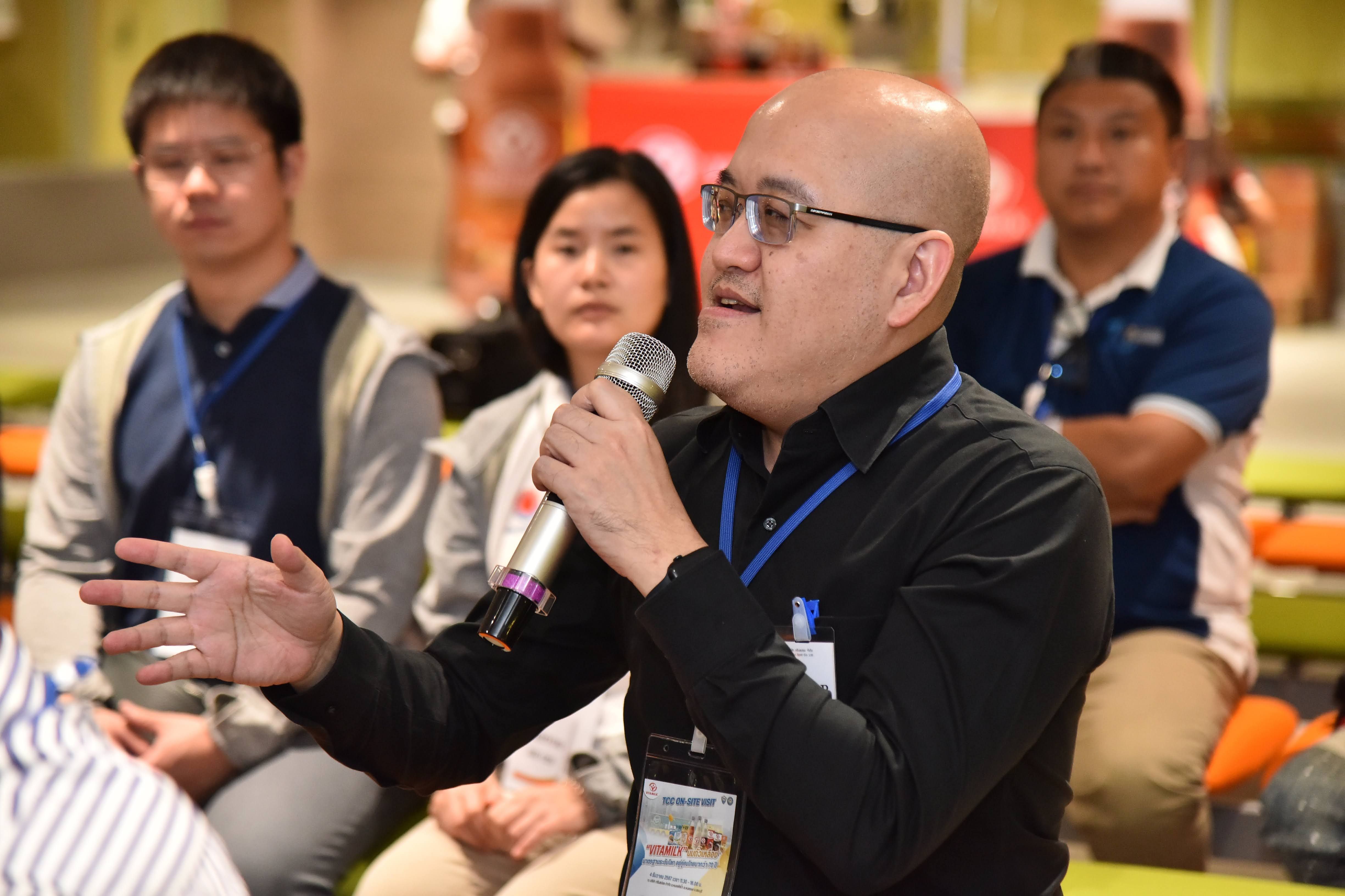 A bald man with glasses speaks into a microphone, gesturing with his hand.