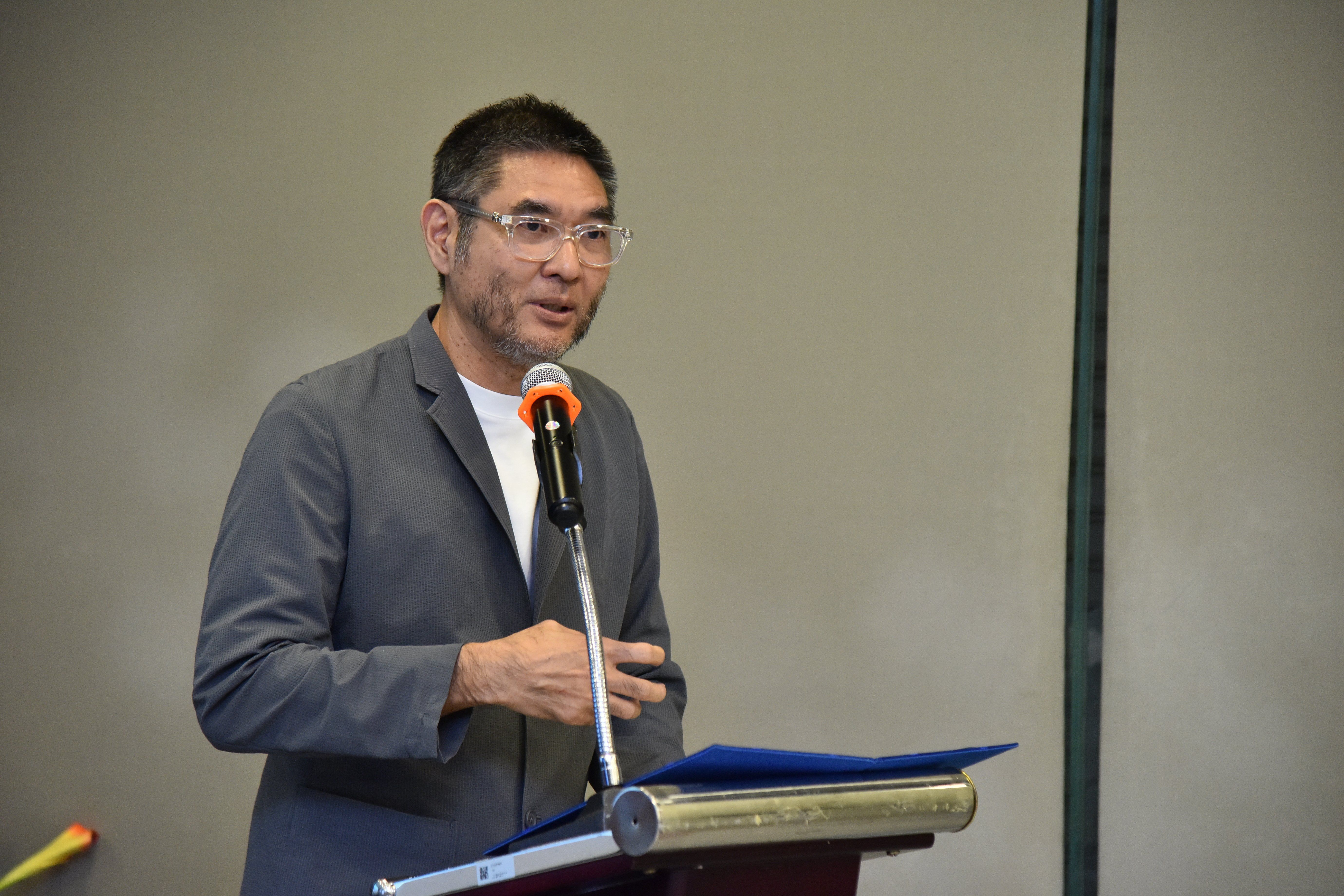 A man with glasses and a grey jacket speaks into a microphone at a podium.