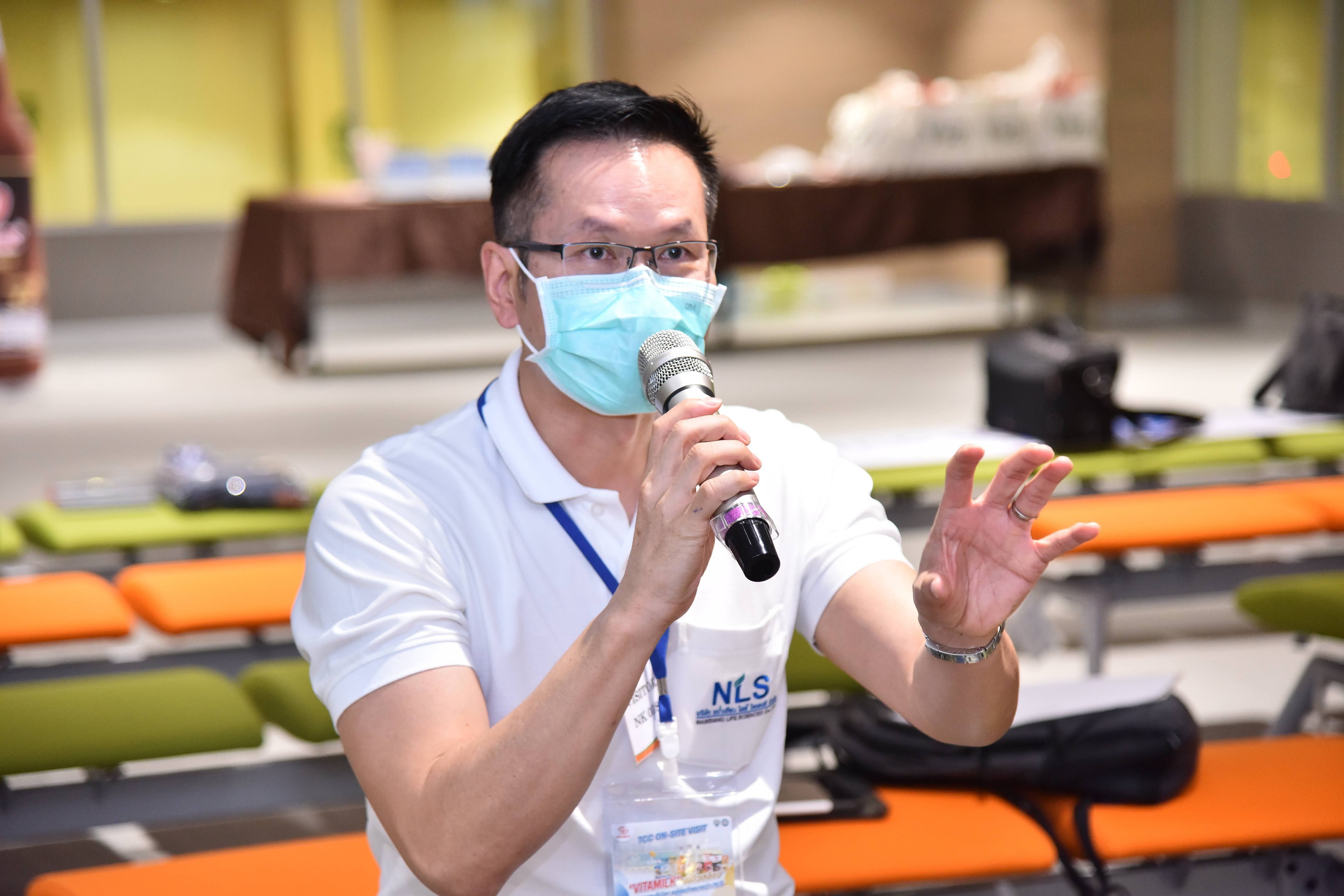 Man in mask and glasses speaks into a microphone, gesturing with his hand at a meeting.